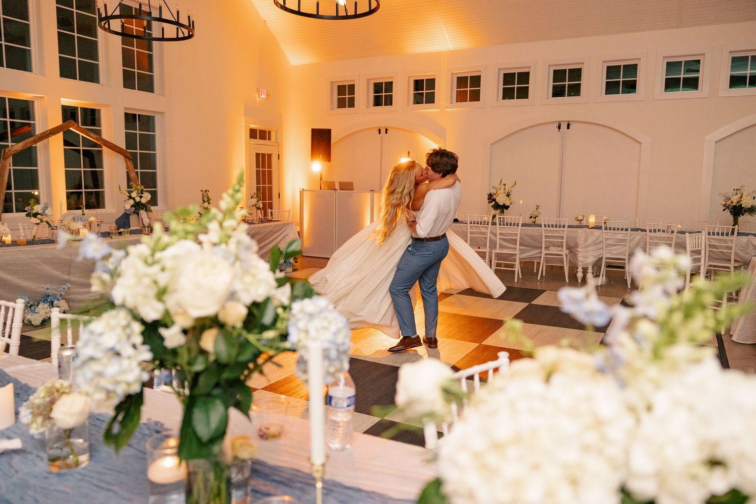 Bride and groom share first dance in elegant white barn wedding venue adorned with white floral centerpieces.