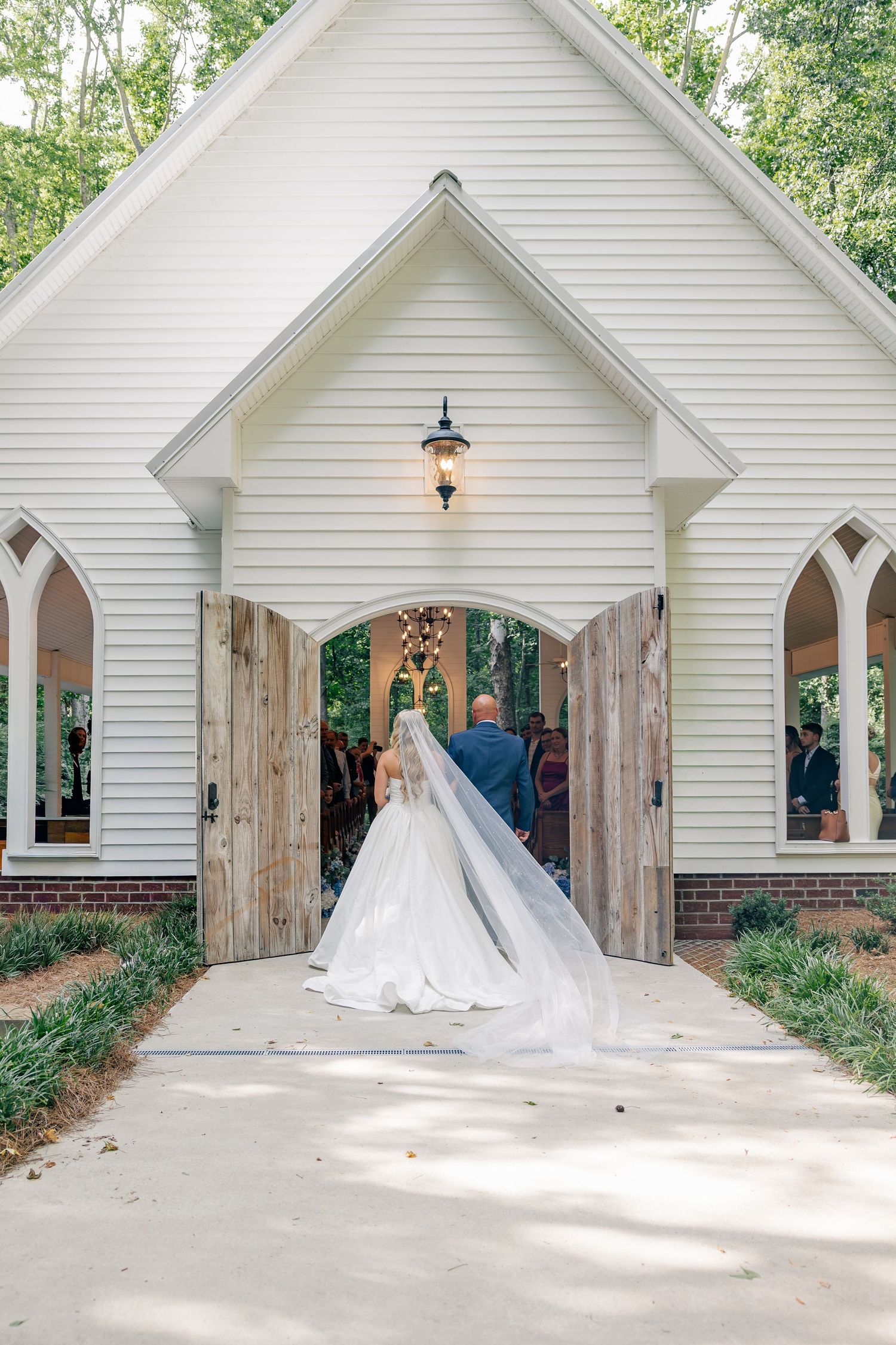 Bride in flowing gown and veil entering white chapel through rustic wooden doors for ceremony.