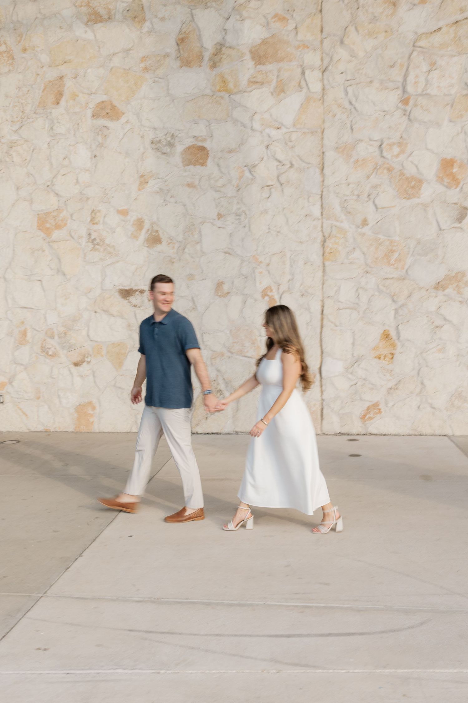 A couple in coordinating white and blue outfits dances playfully against a textured stone wall.