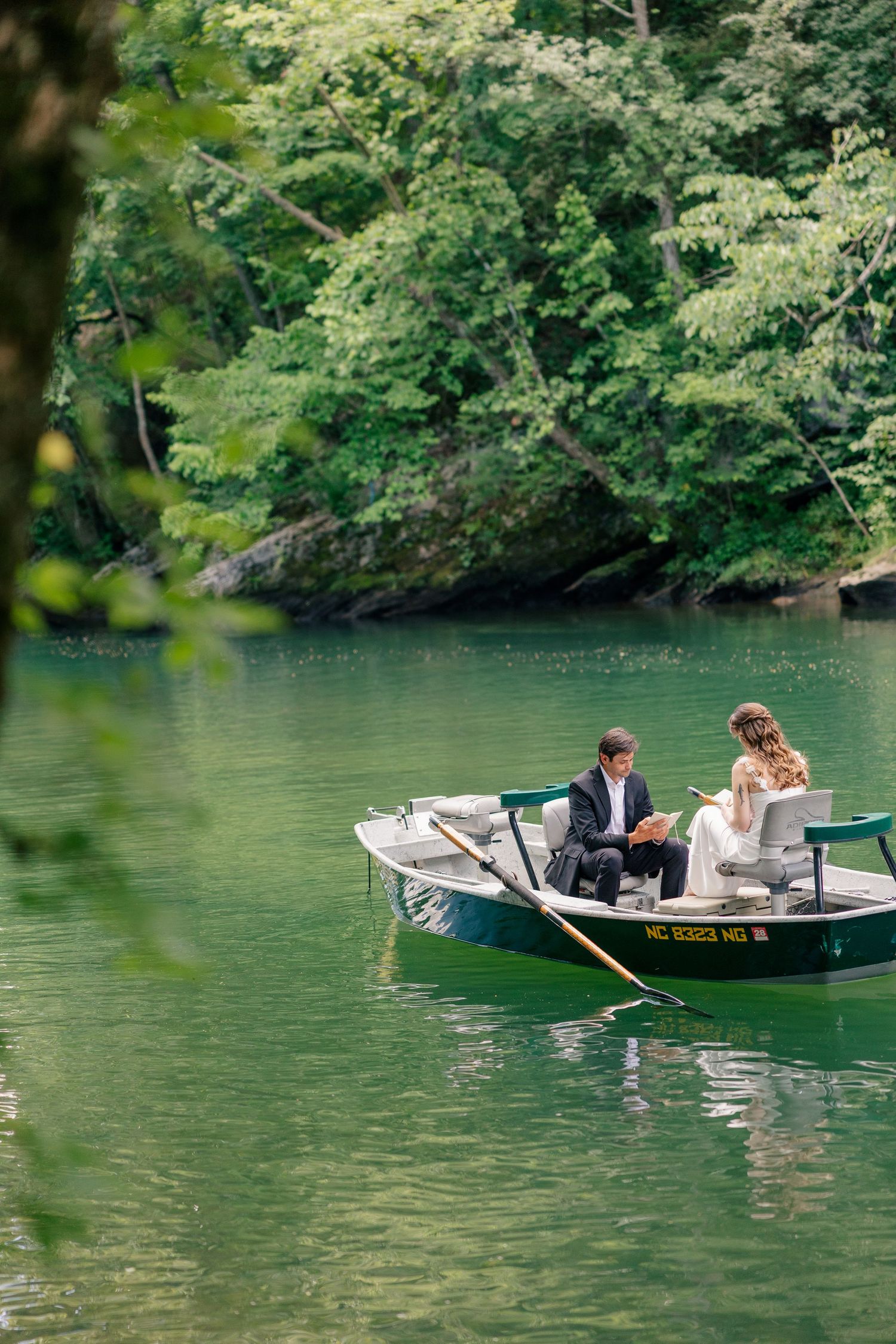 Newlywed couple shares romantic rowboat ride on a serene green lake surrounded by trees.