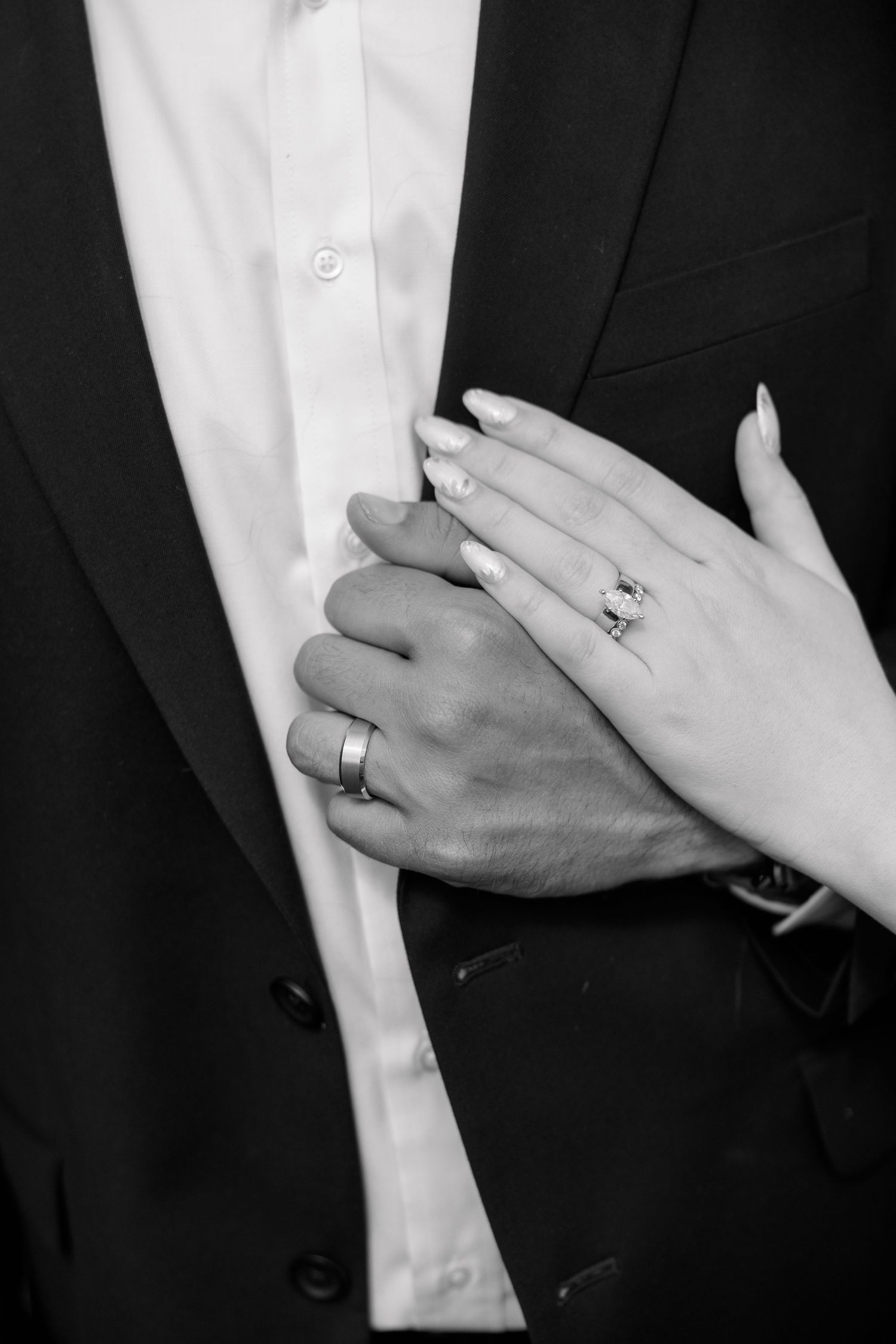 Black and white close-up of bride and groom holding hands showing wedding rings against dark suit.