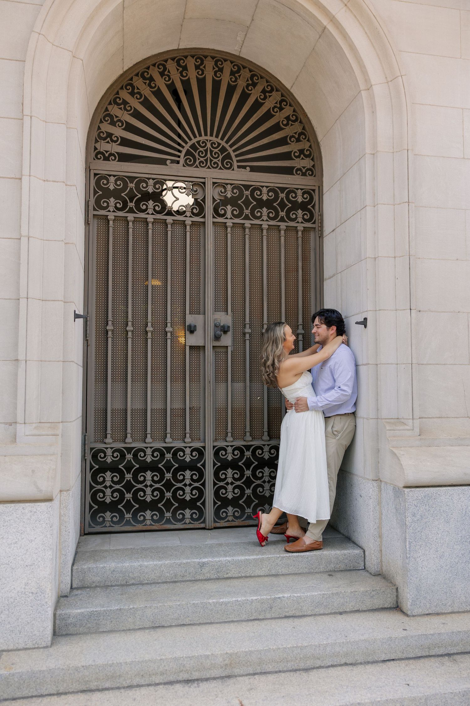 Couple embracing in front of ornate iron gate doorway, woman in white dress and red heels.