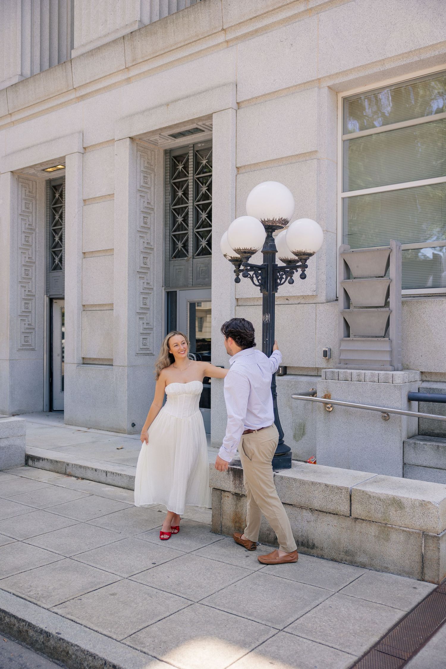 Couple in white dress and khakis dancing by ornate street lamp outside classical stone building.