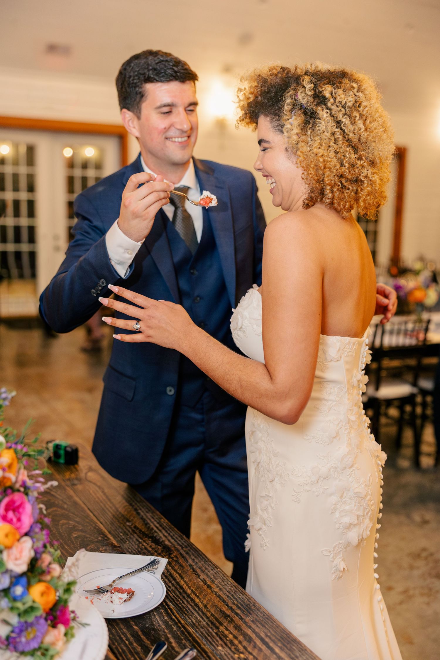 Bride and groom laugh joyfully while cutting wedding cake indoors, surrounded by colorful floral arrangements.