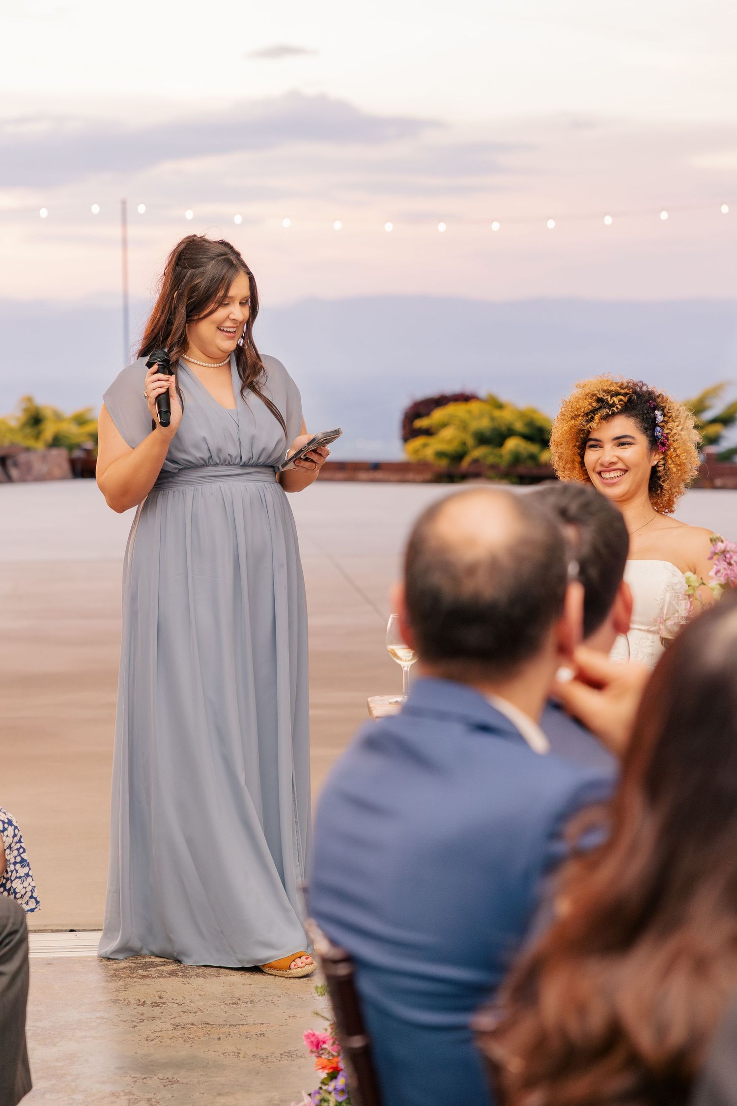 Bridesmaid in blue gown gives speech at outdoor wedding ceremony while bride smiles nearby.