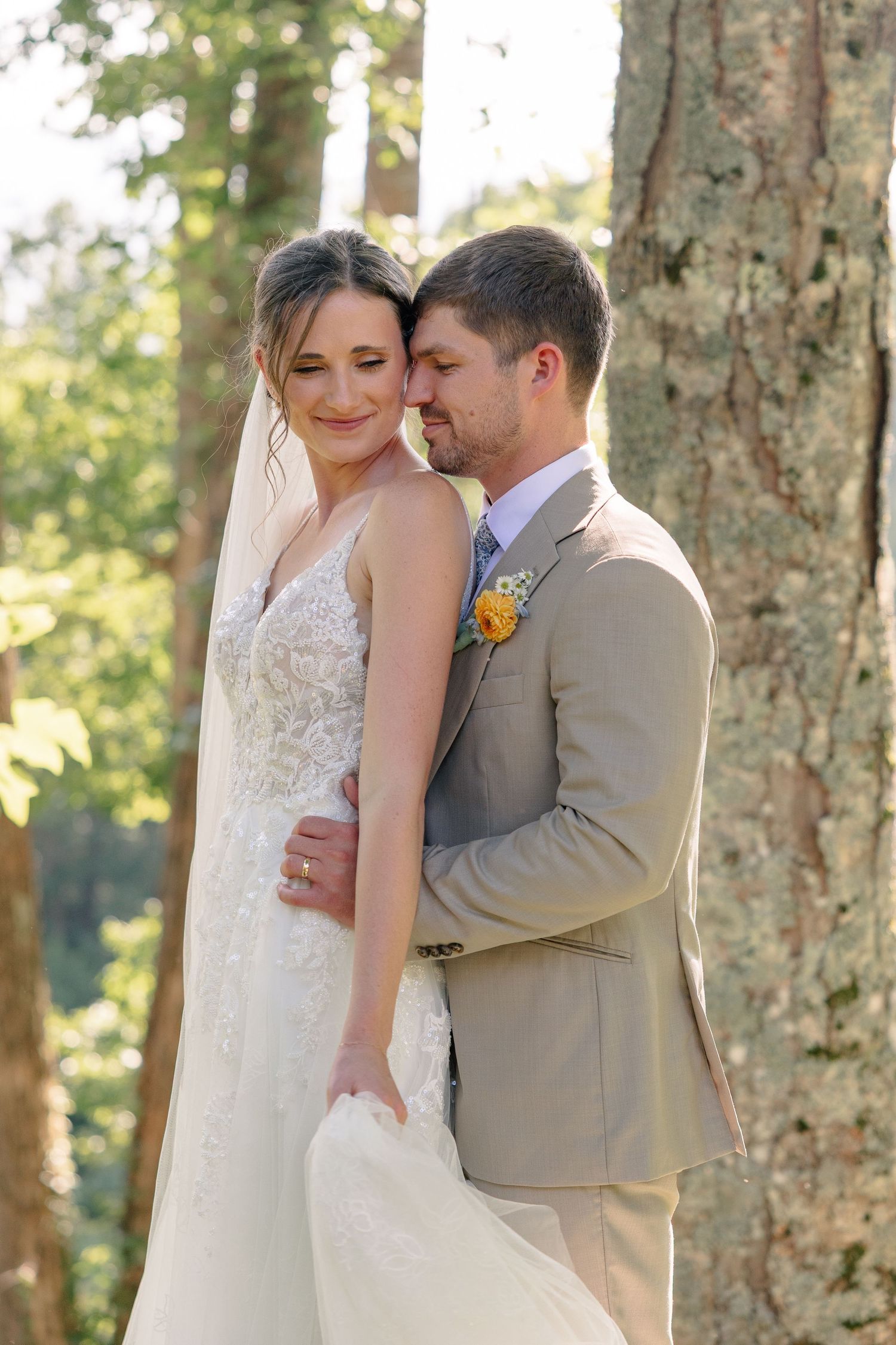 A bride and groom share a tender embrace outdoors among trees, she in a lace gown with veil, he in a tan suit.