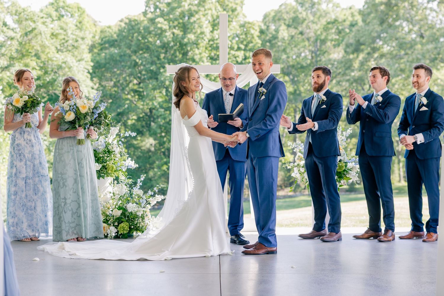 Bride and groom exchange vows at outdoor pavilion ceremony surrounded by bridesmaids and groomsmen in blue.