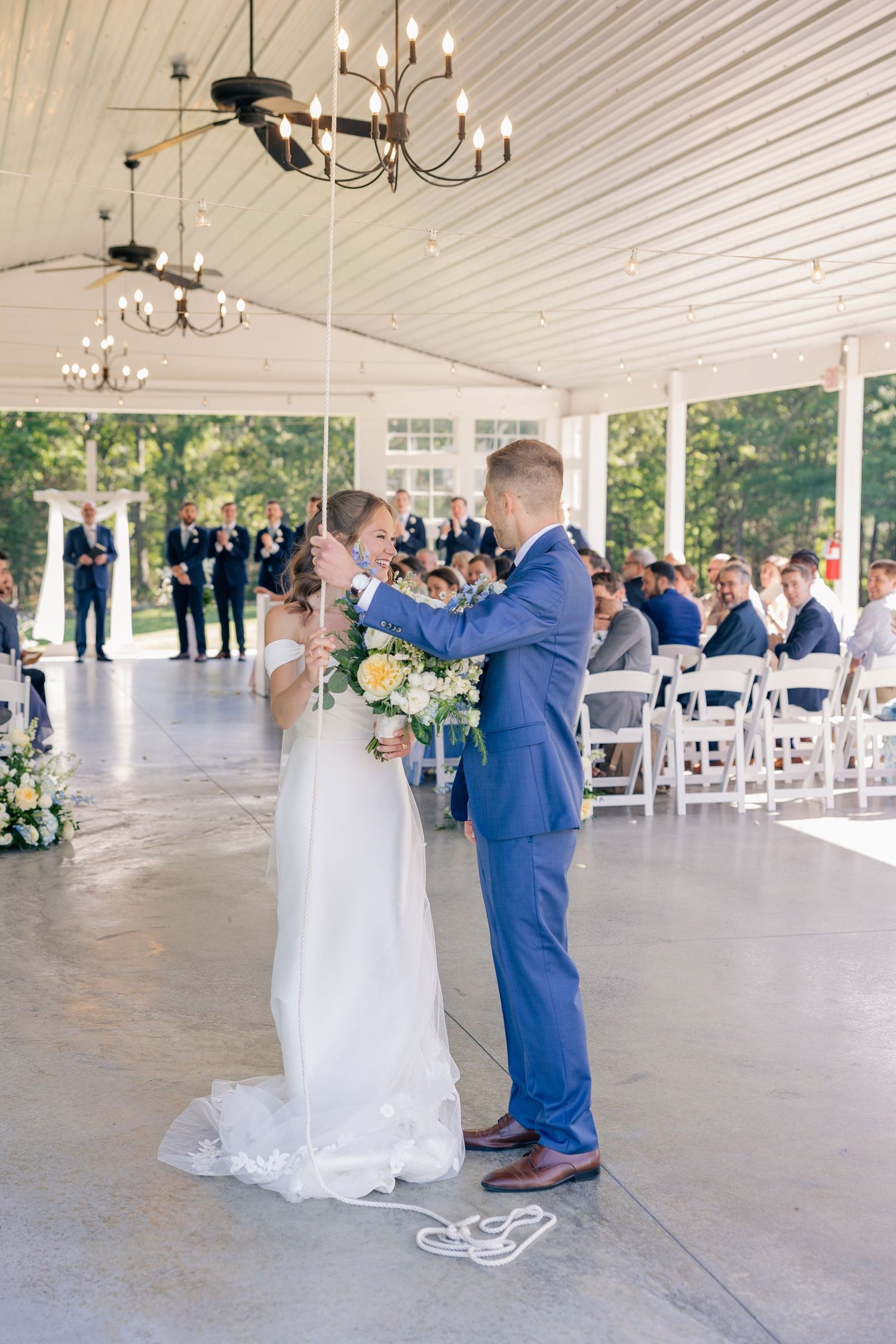 Bride and groom share first look moment holding bouquet under chandelier in white open-air wedding pavilion.