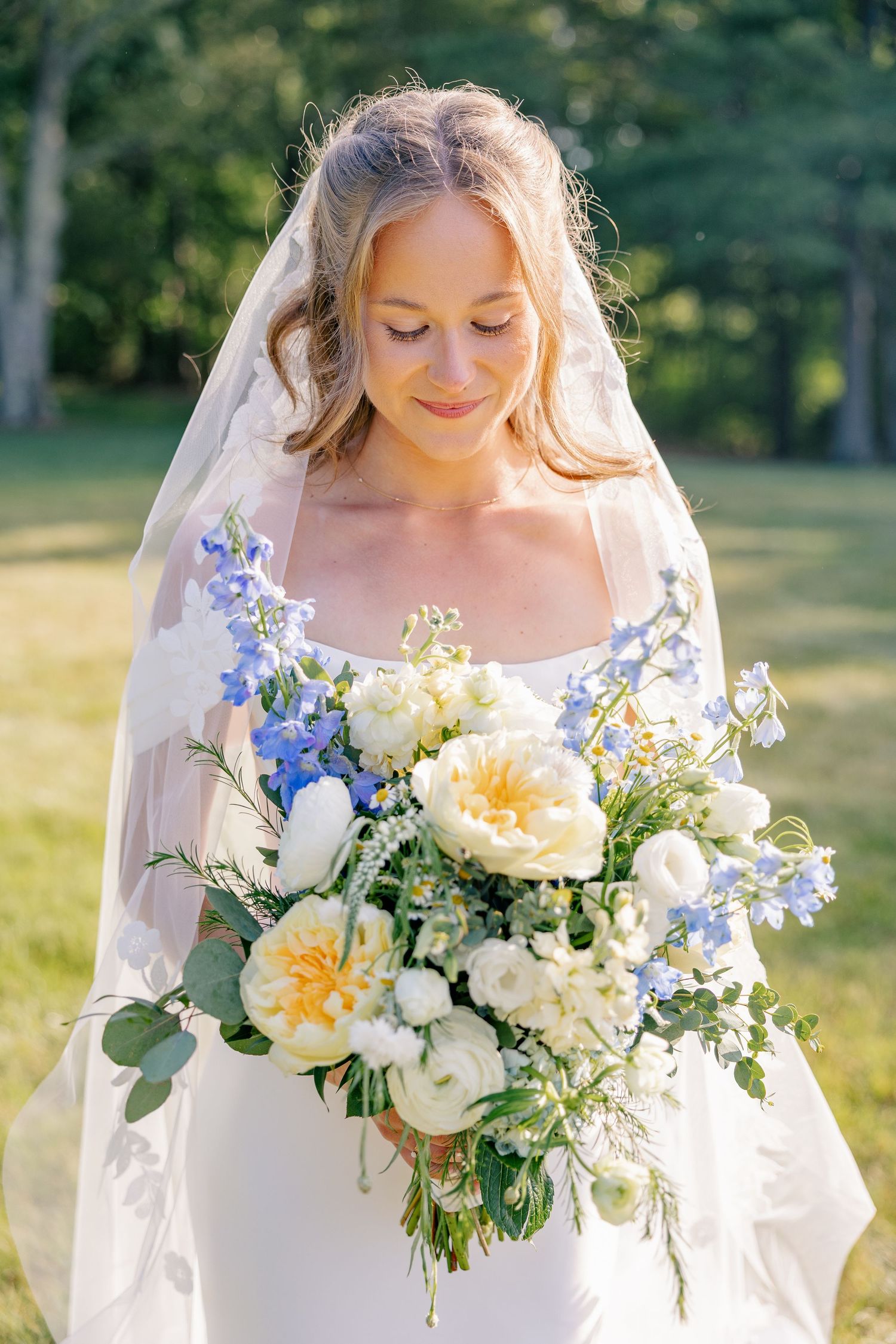 Beautiful bride in white veil gazes down at lush bouquet of blue delphiniums and cream peonies.