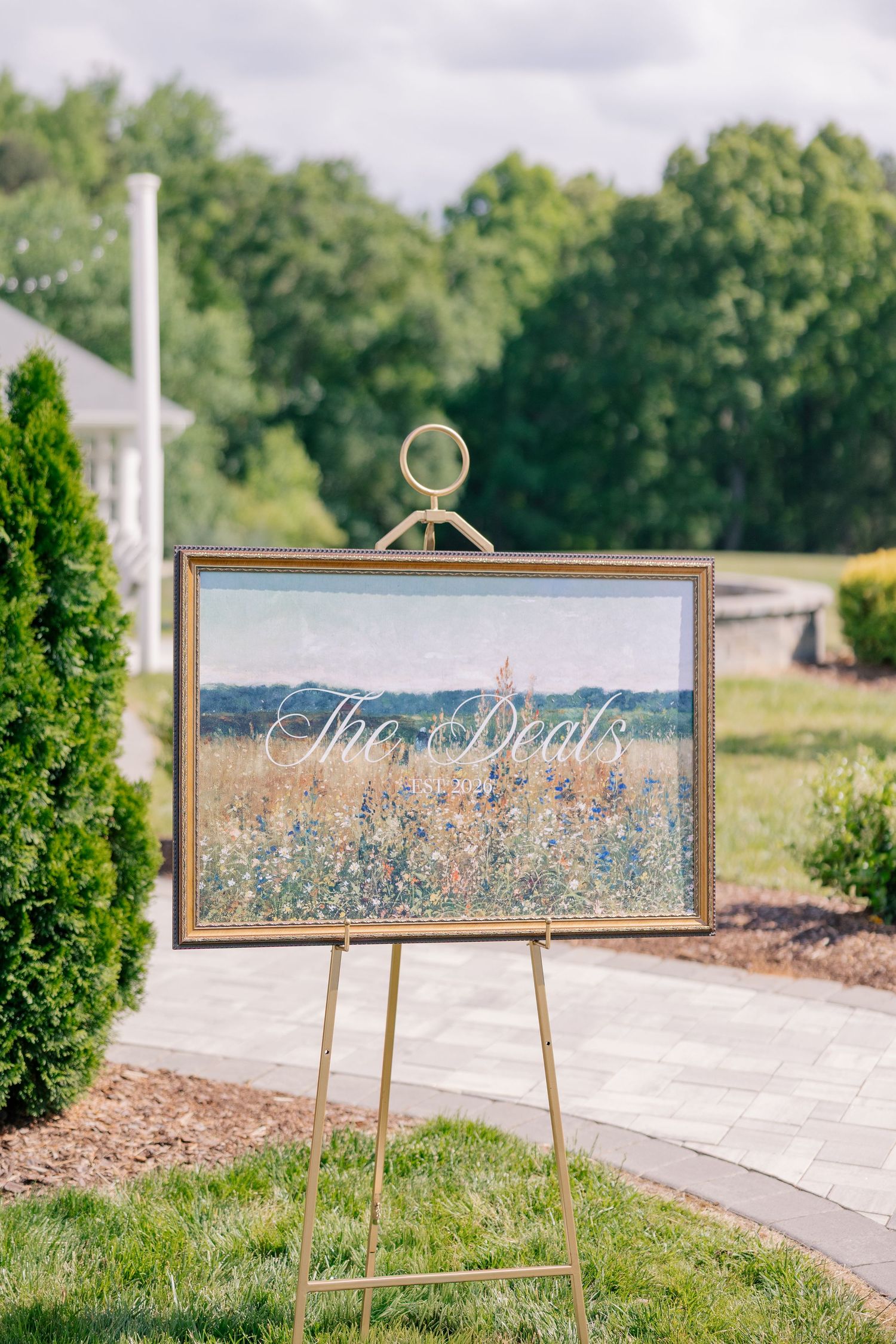 Gold-framed wildflower field painting on easel reads The Deal at outdoor wedding venue.