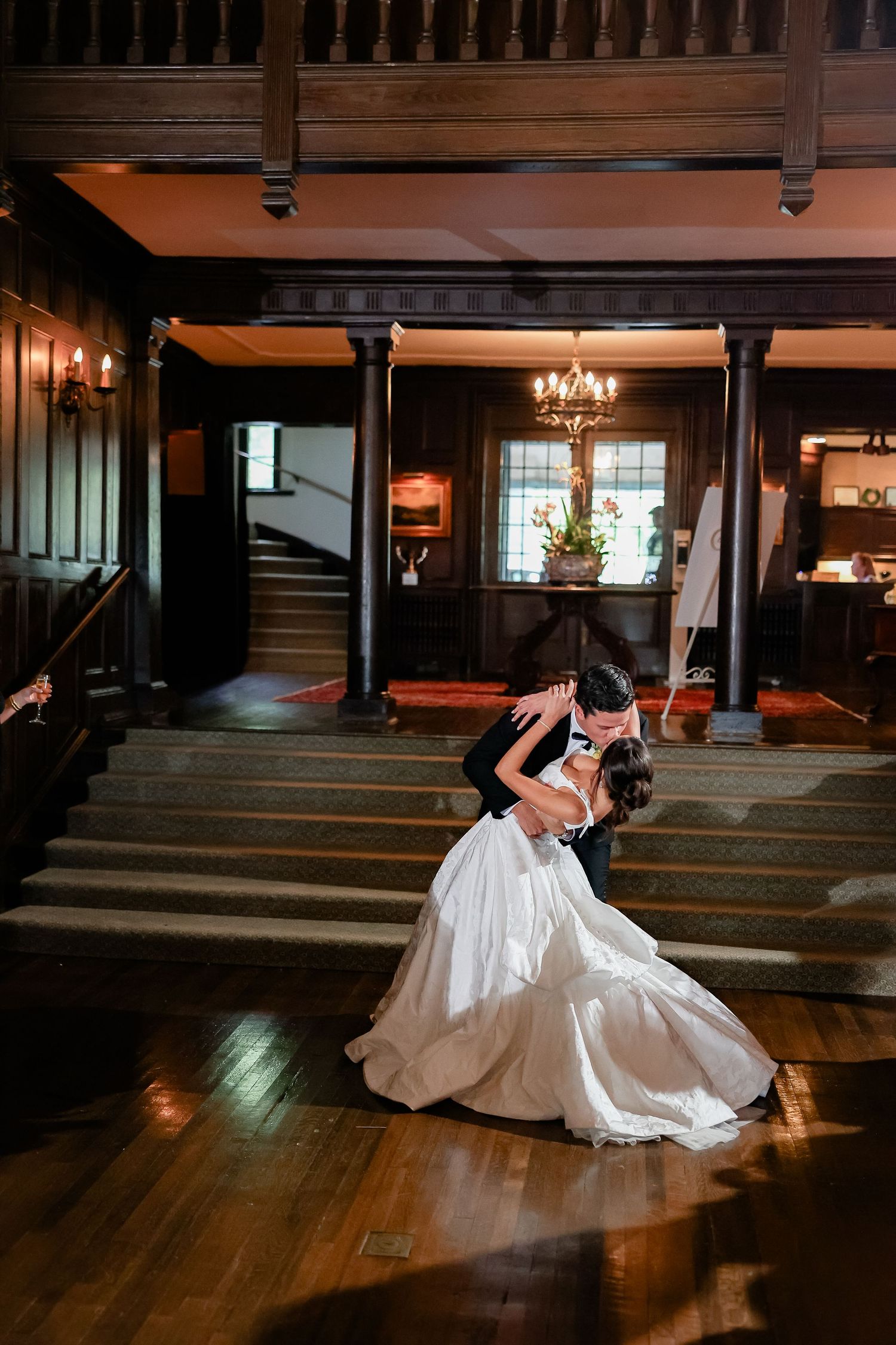 Bride and groom share a romantic dip kiss on a wooden dance floor in an elegant wood-paneled ballroom.