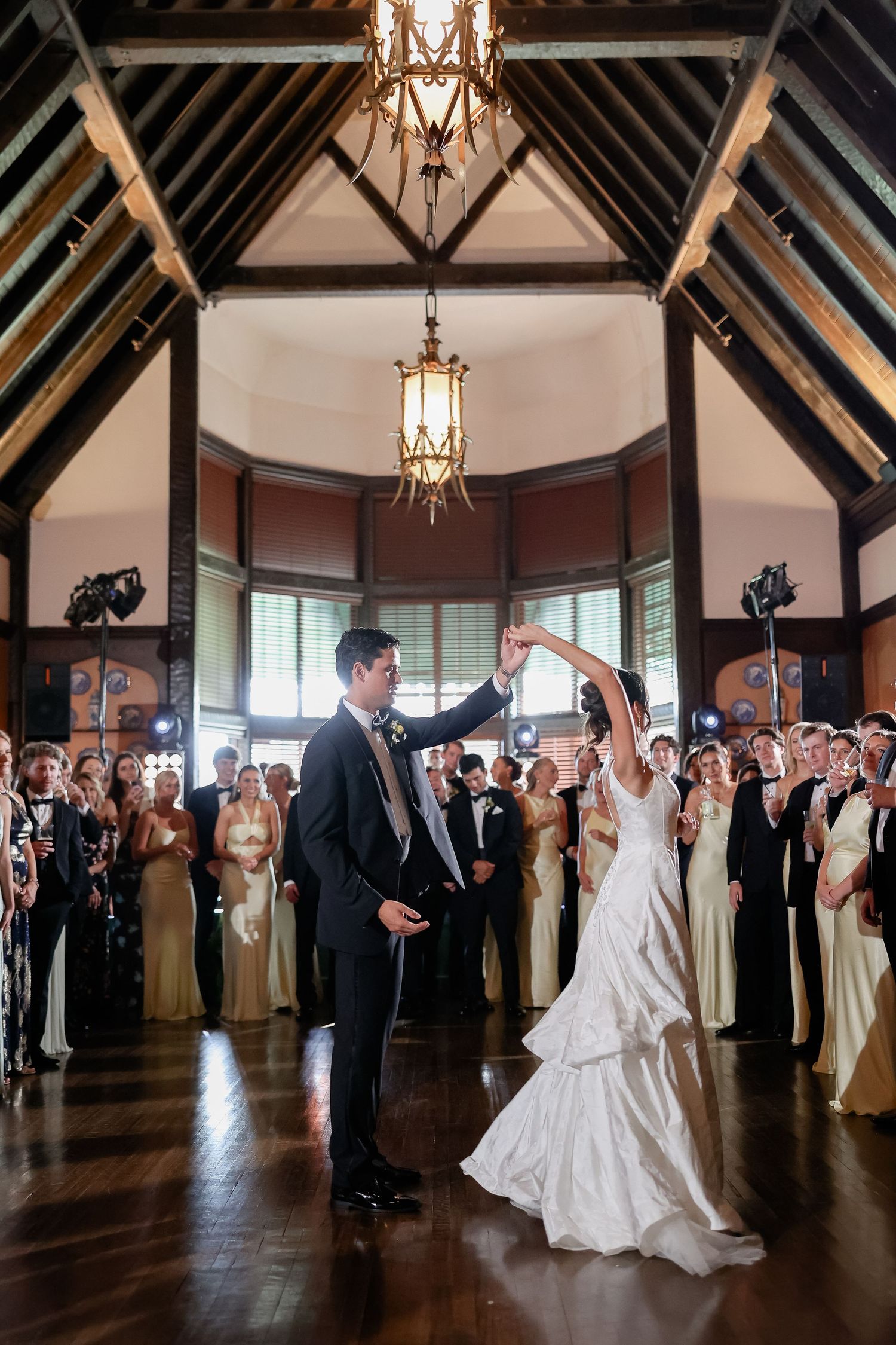 Bride and groom first dance under a chandelier in an elegant wood-beamed ballroom surrounded by wedding party guests.