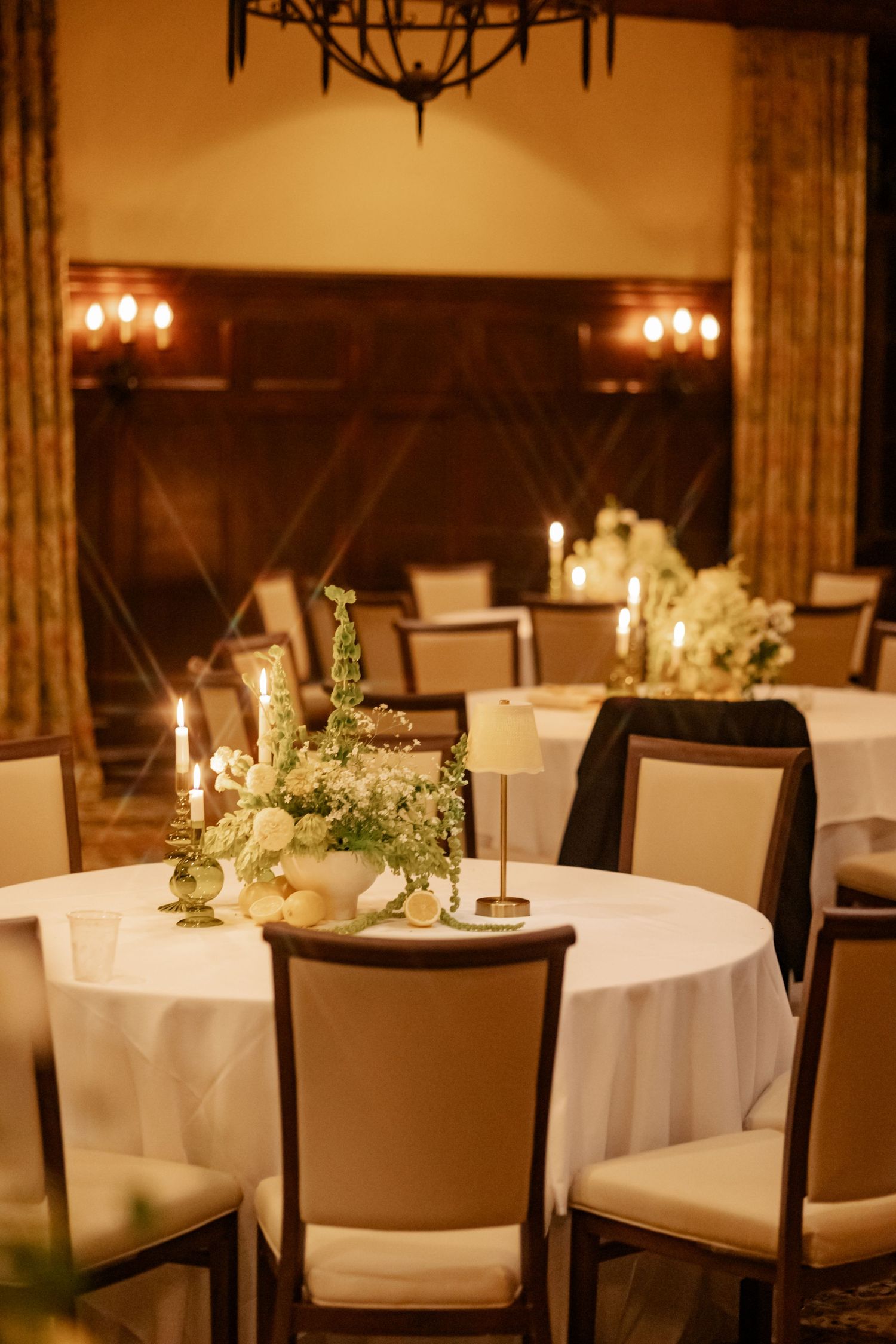 Elegant wedding reception with round tables, white linens, candlelight, and floral centerpieces in a warmly lit ballroom.