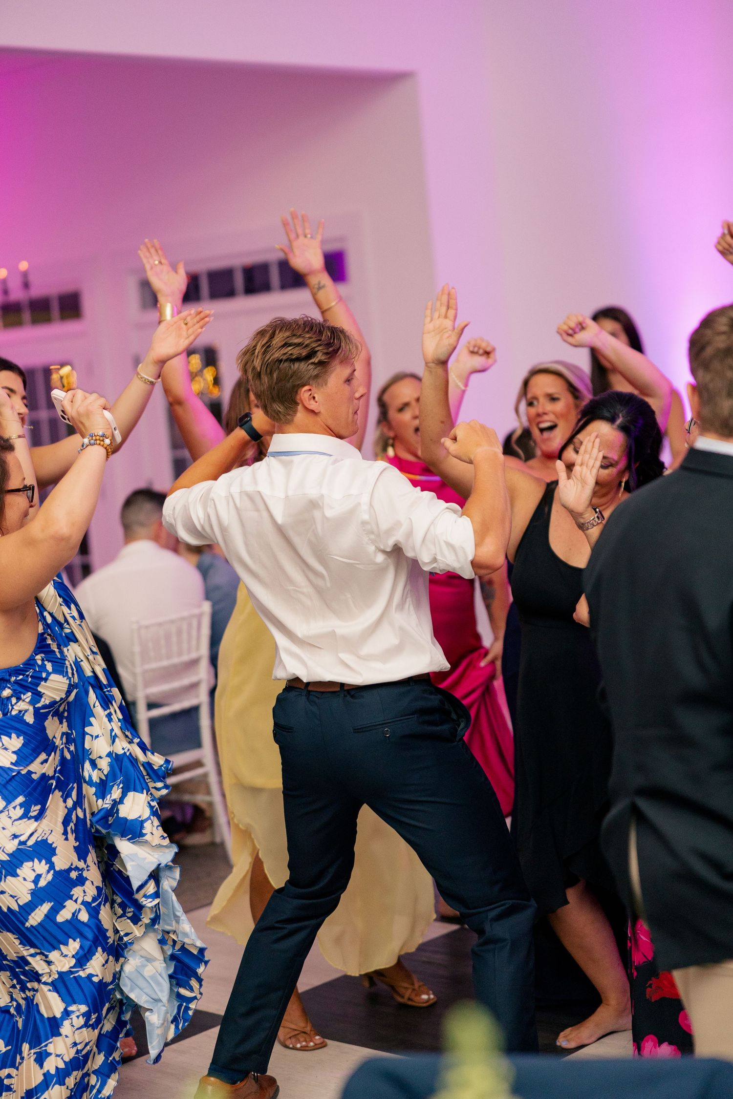 Guests dancing with hands raised at a lively wedding reception with pink ambient lighting.