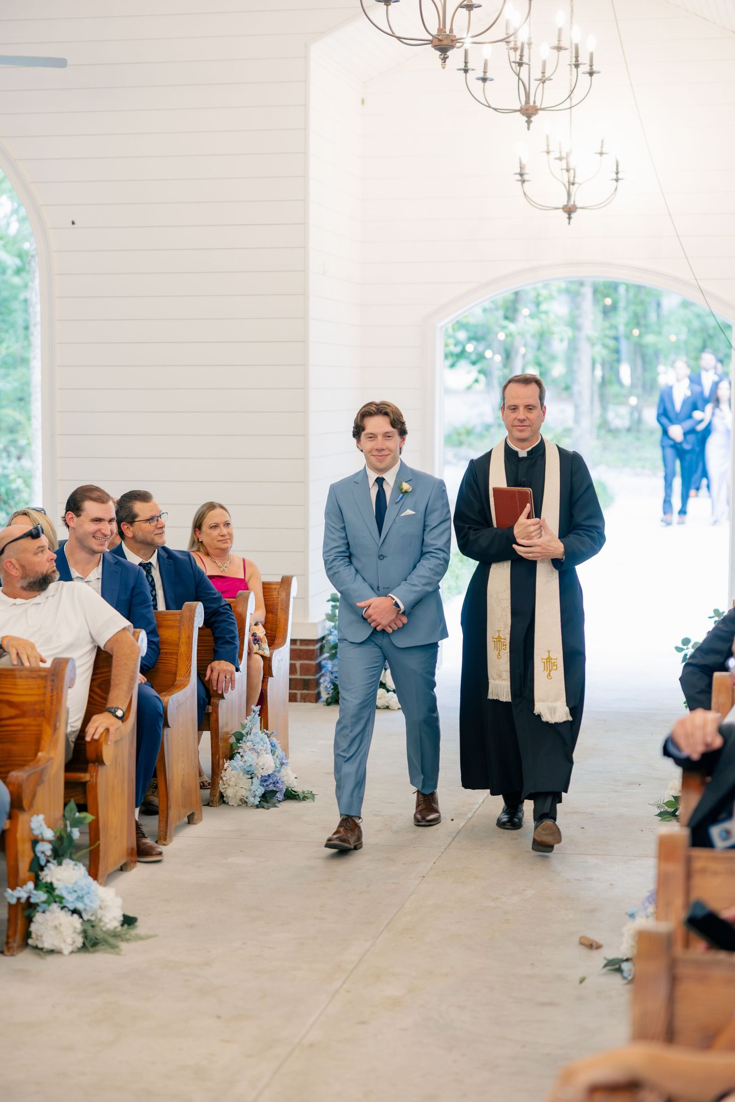 Groom walking down aisle with officiant in elegant white chapel ceremony.