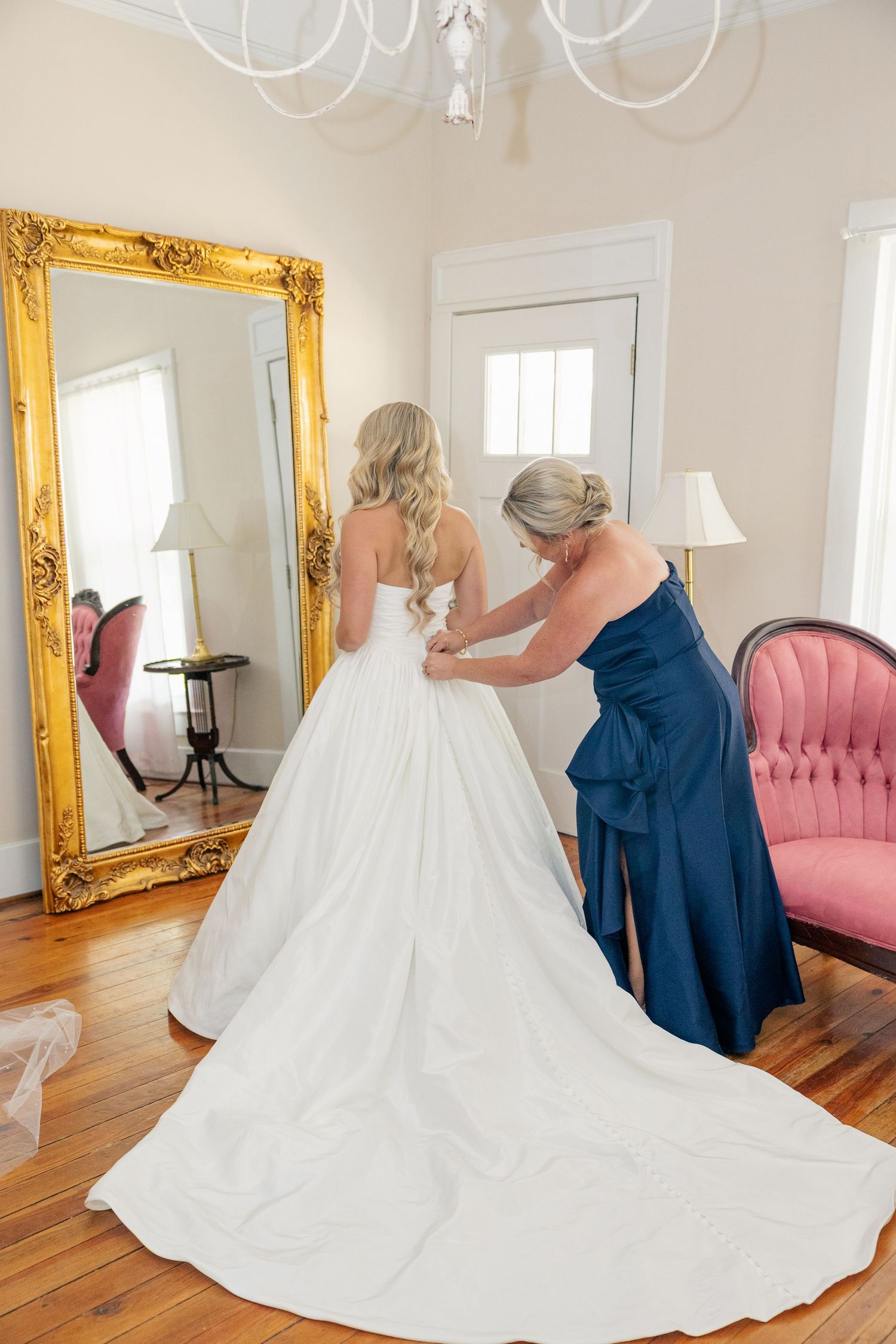Woman in navy dress helping bride button up her elegant white ball gown wedding dress.