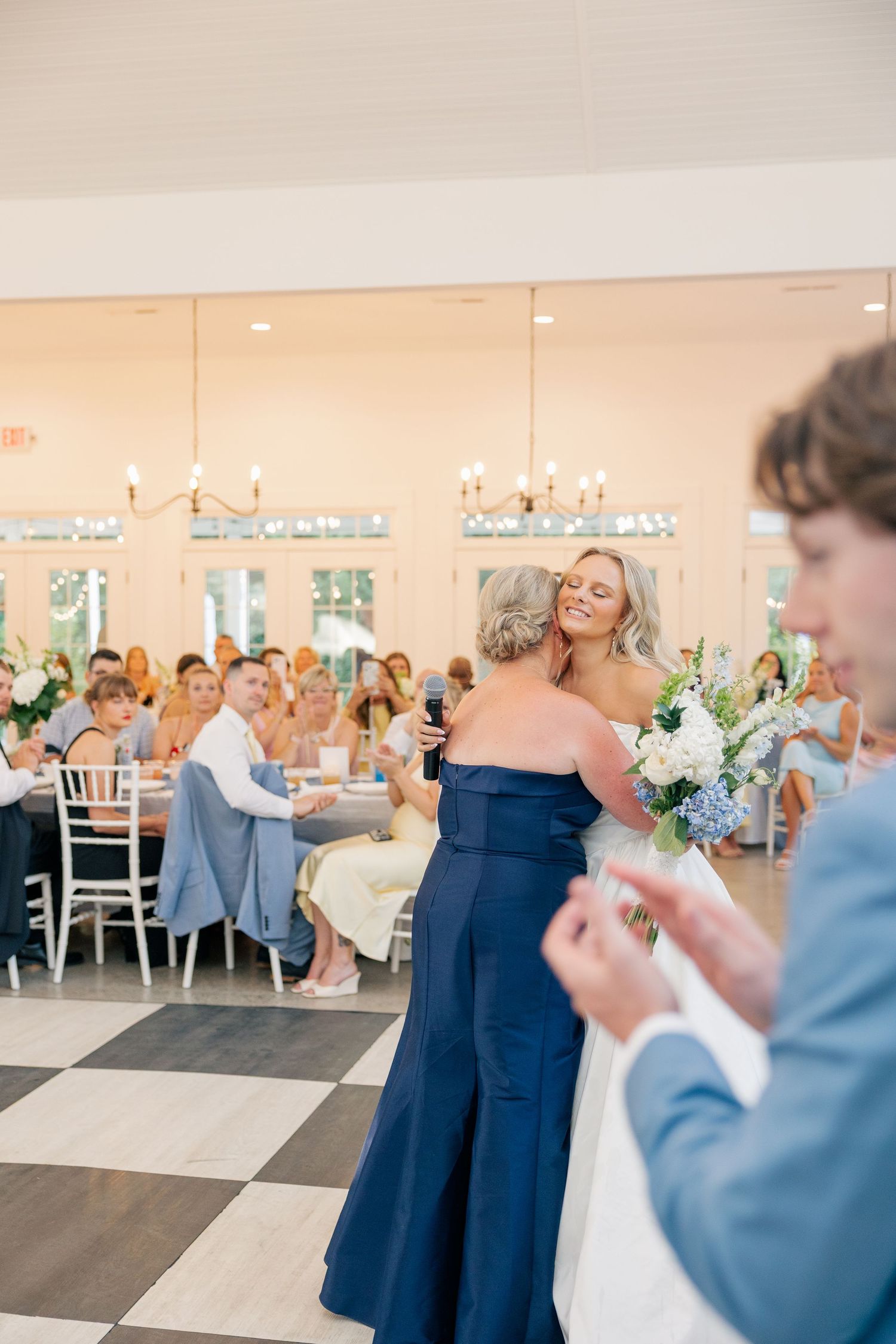 Wedding guests gathered in elegant reception hall as bridesmaid in navy gown embraces woman during toast.