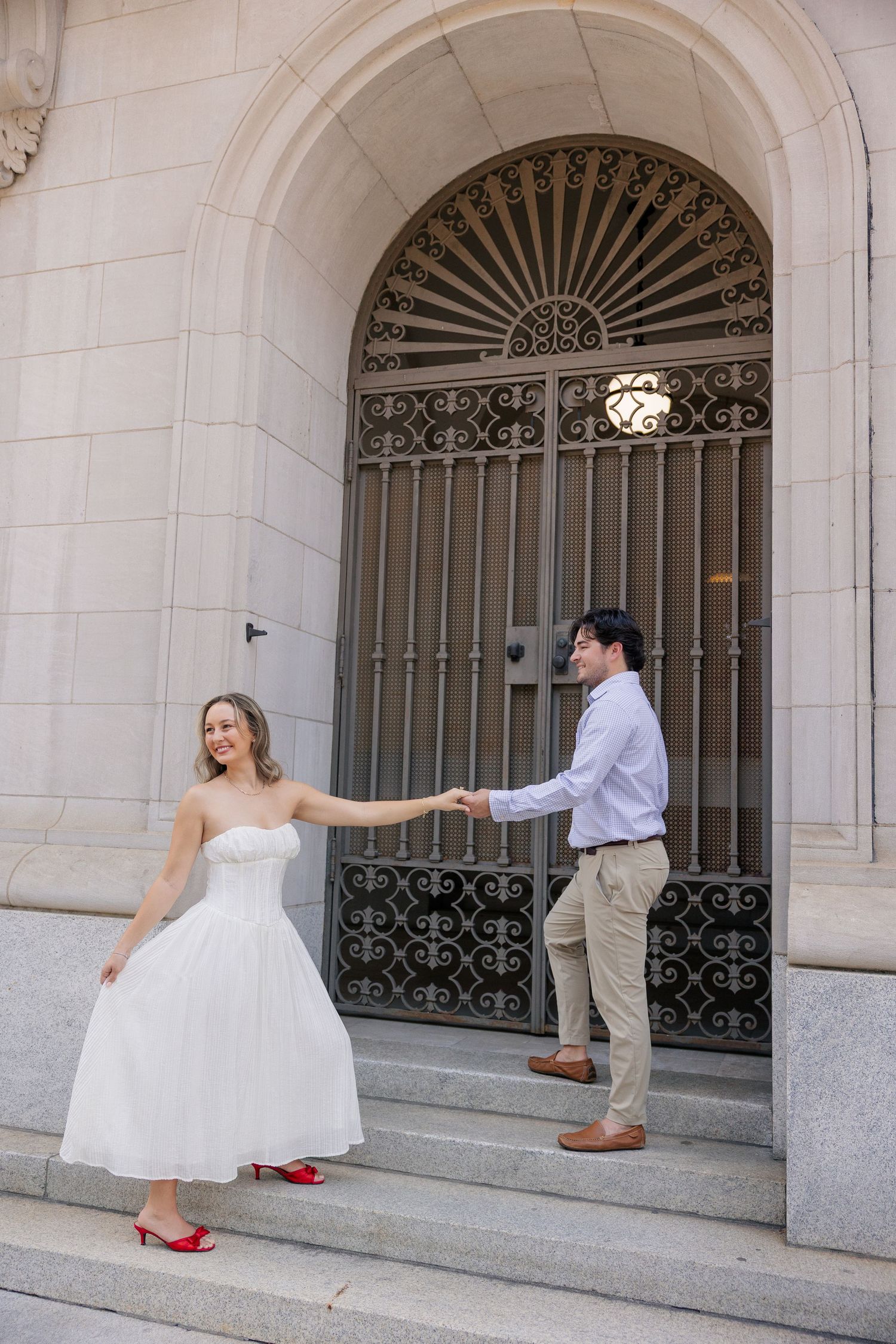 Bride in white dress with red heels holds grooms hand on steps of elegant stone building with ornate iron door.