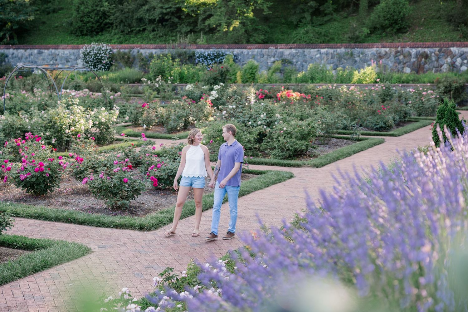 Two people stroll along a garden path surrounded by lavender and colorful summer blooms in a formal garden setting.