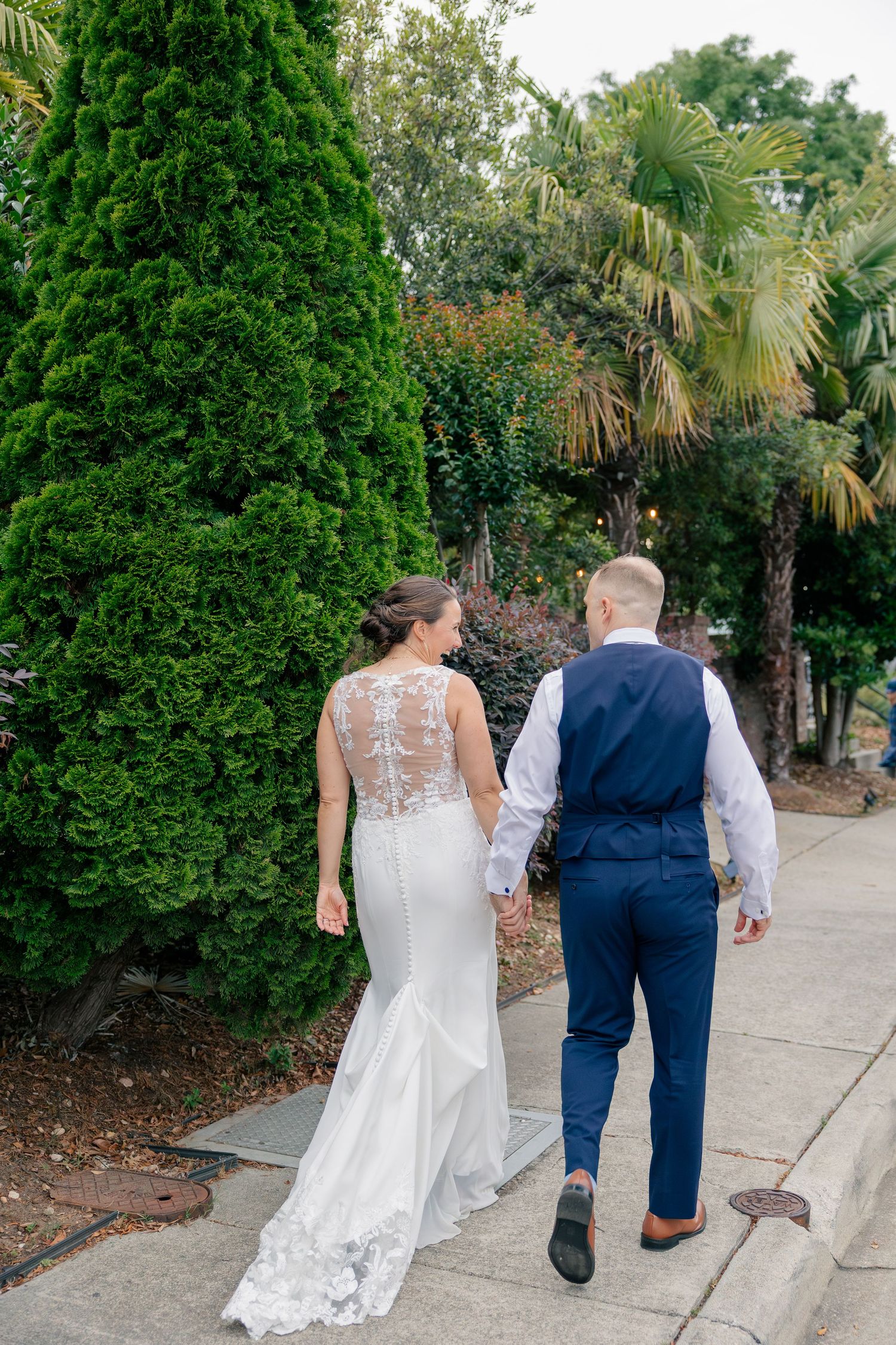 Bride and groom walk hand in hand past lush greenery and palm trees after wedding ceremony.