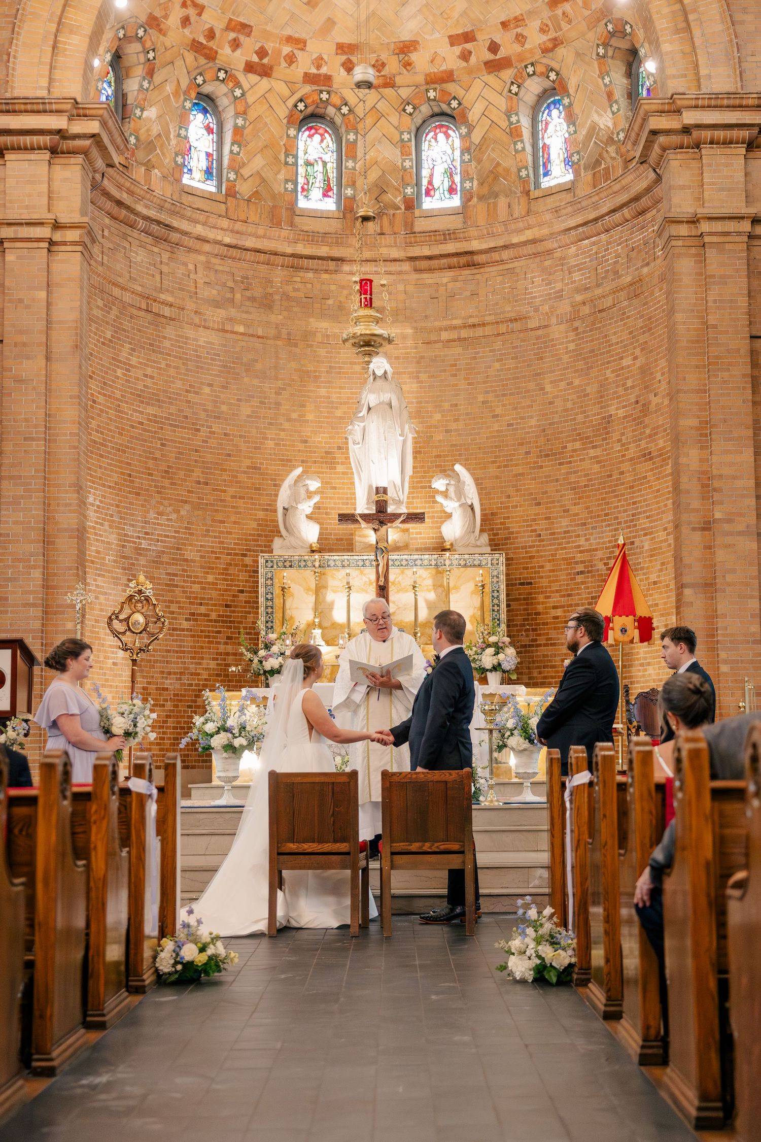 Bride and groom exchanging vows at ornate brick Catholic church altar with stained glass windows.