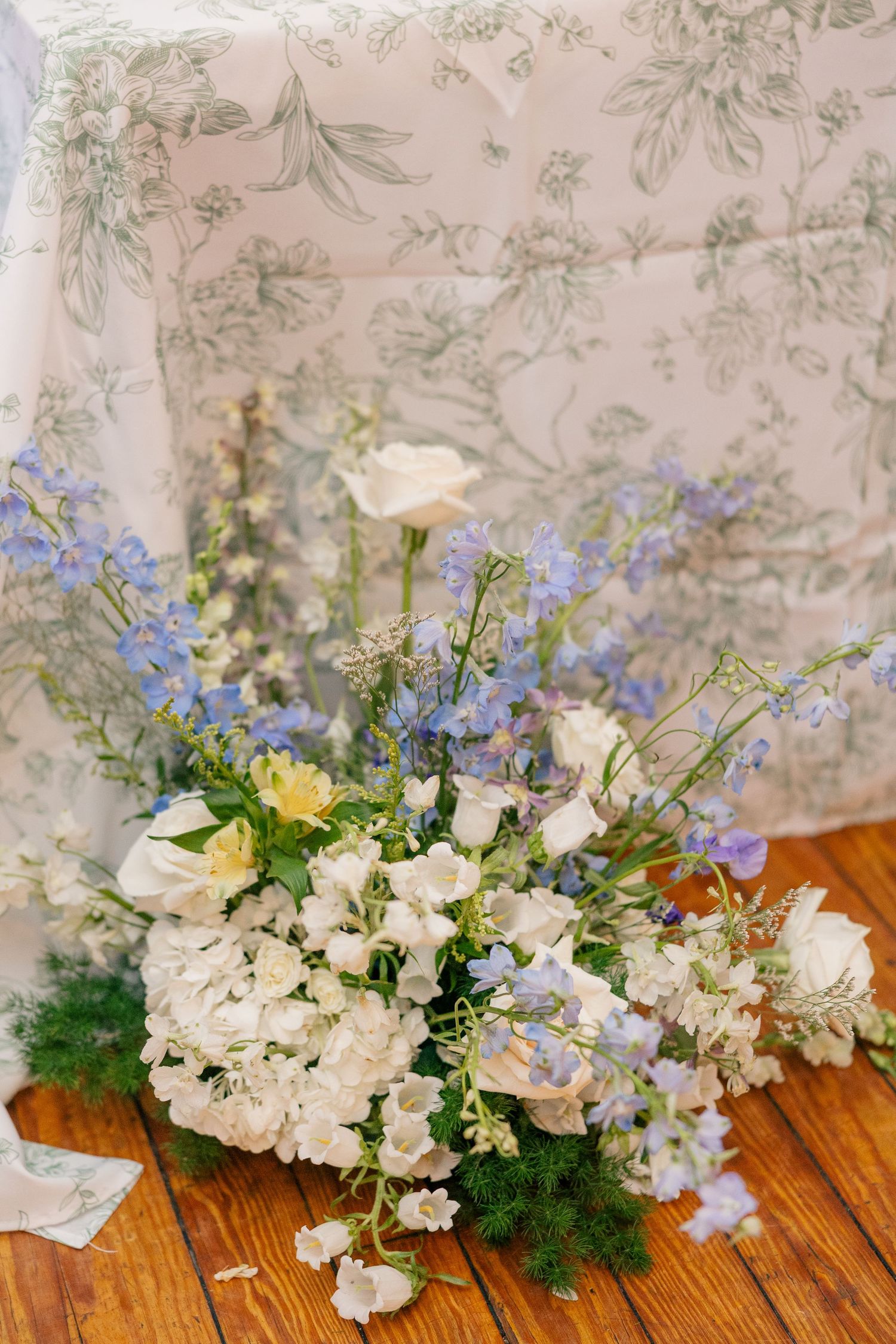 Romantic wedding floral arrangement with white roses, blue delphiniums, and greenery on a wooden floor beside a floral tablecloth.