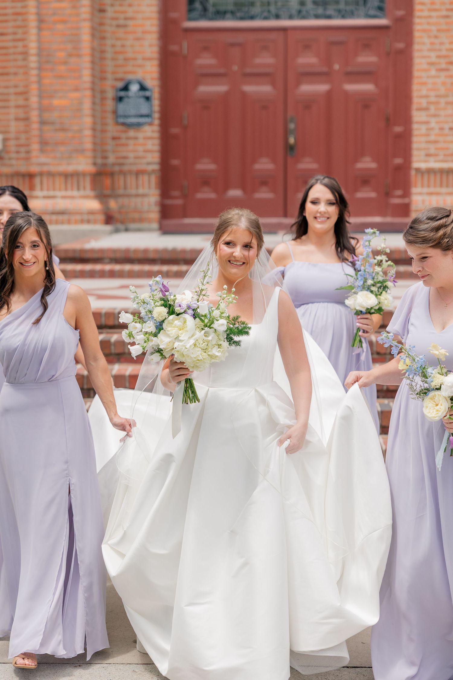 Bride in white ball gown walks with bridesmaids in lavender dresses outside red-doored brick church.