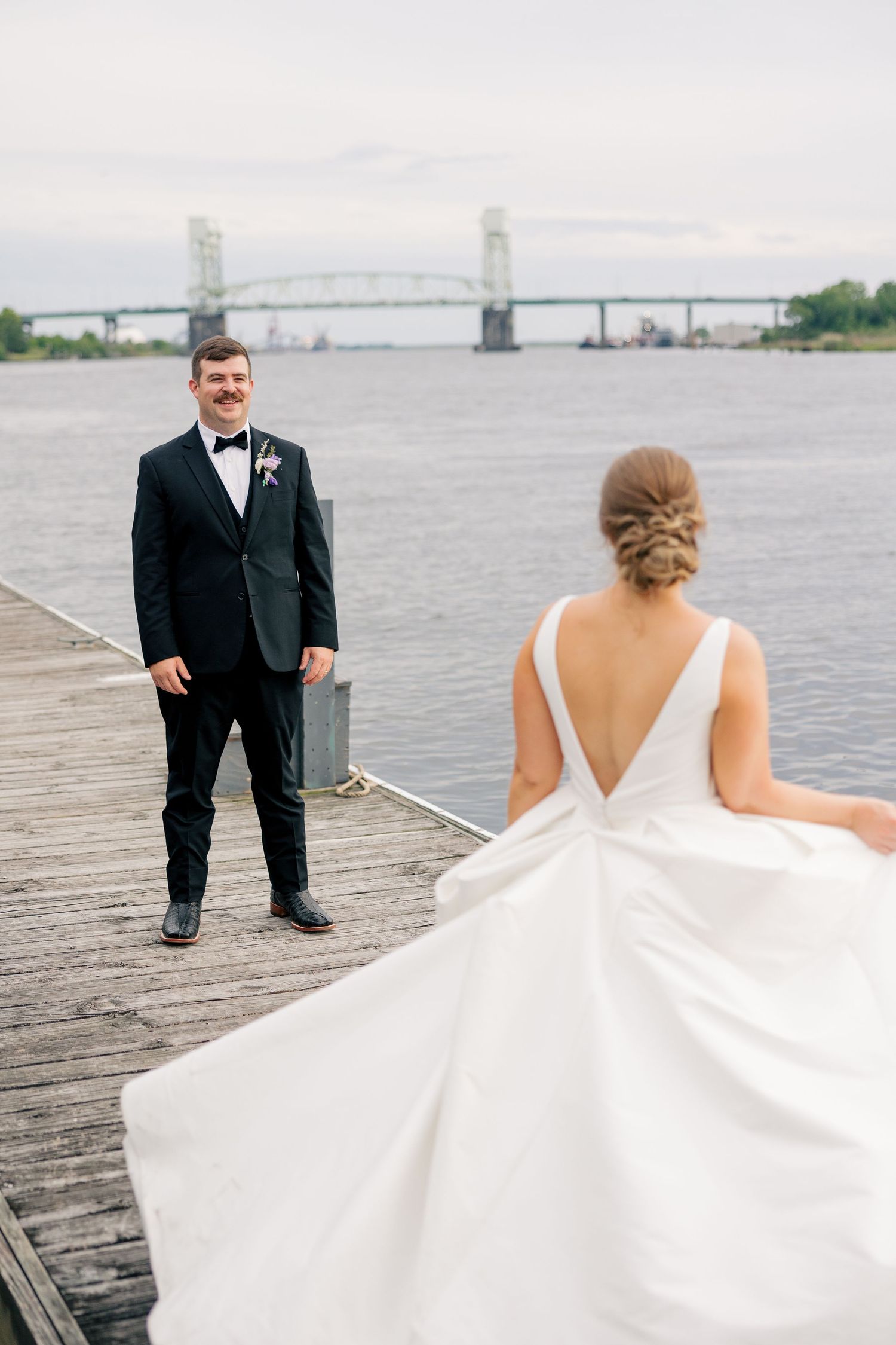 Groom in black tuxedo smiles at bride on wooden dock with bridge and river backdrop.