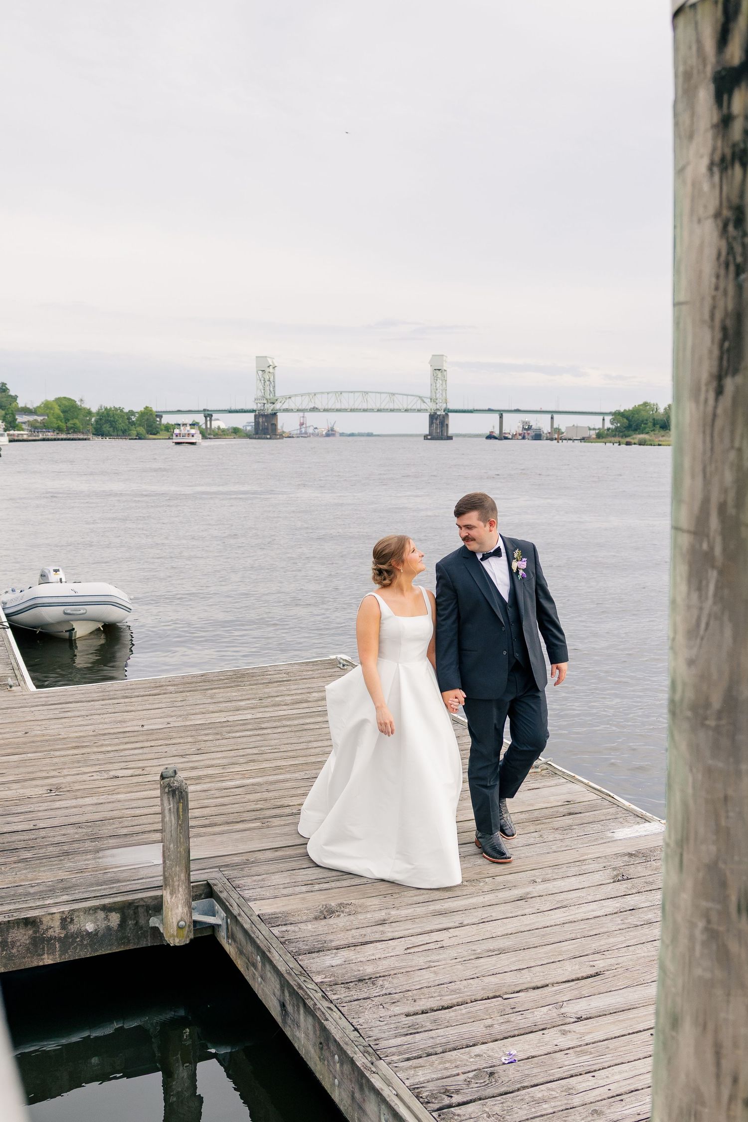 Bride and groom walk hand in hand on a wooden dock with a scenic river bridge backdrop.