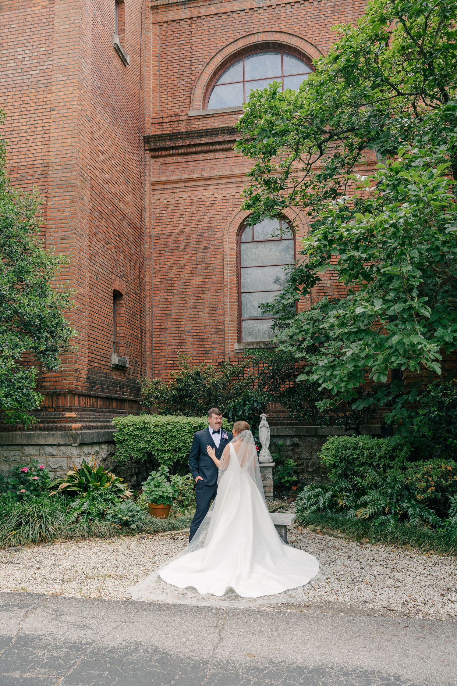 Bride and groom posing romantically outside historic red brick church surrounded by lush greenery.