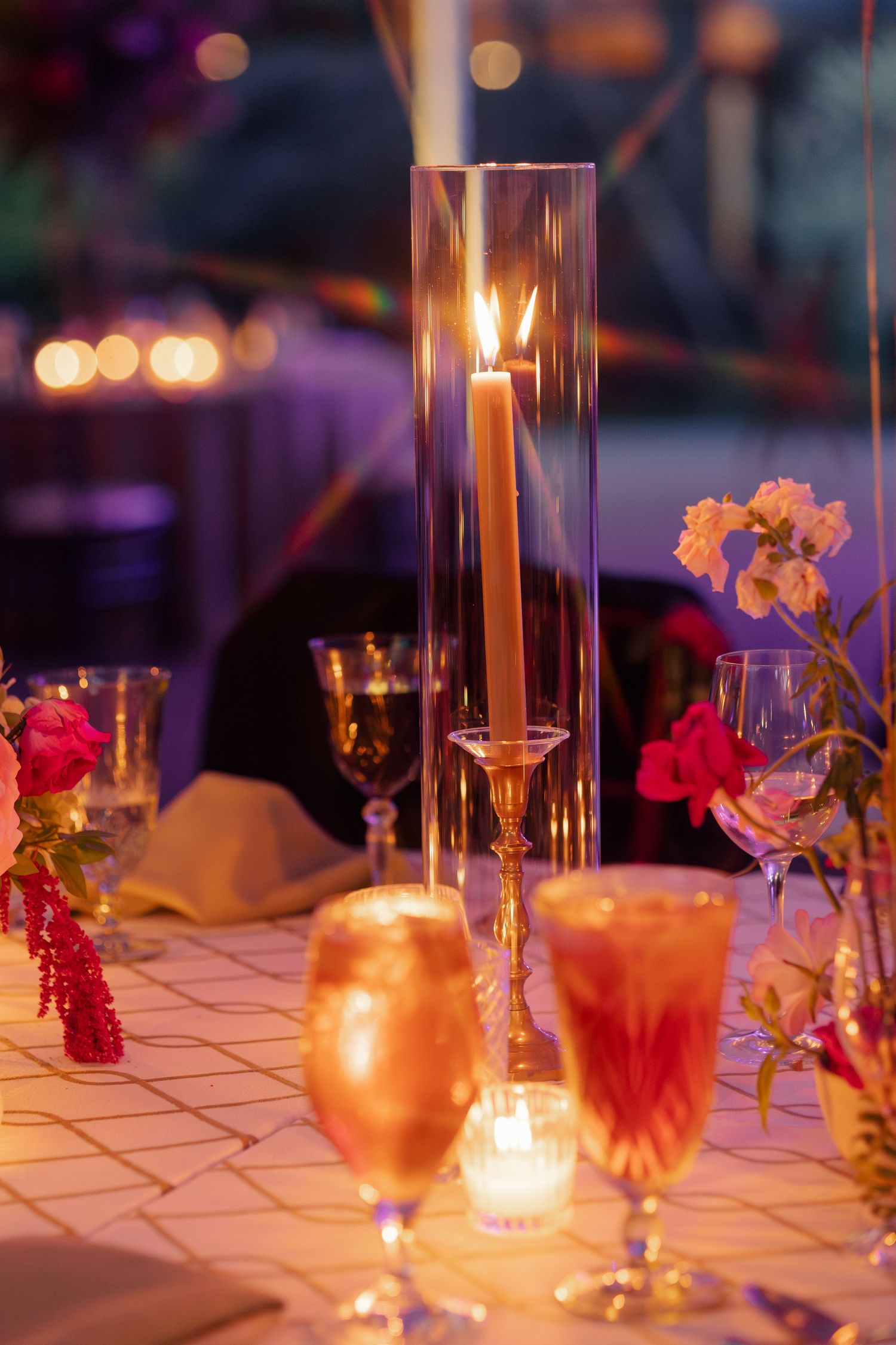 Elegant candlelit reception table with tall glass candle holder, pink florals, and glowing stemware at a romantic evening event.