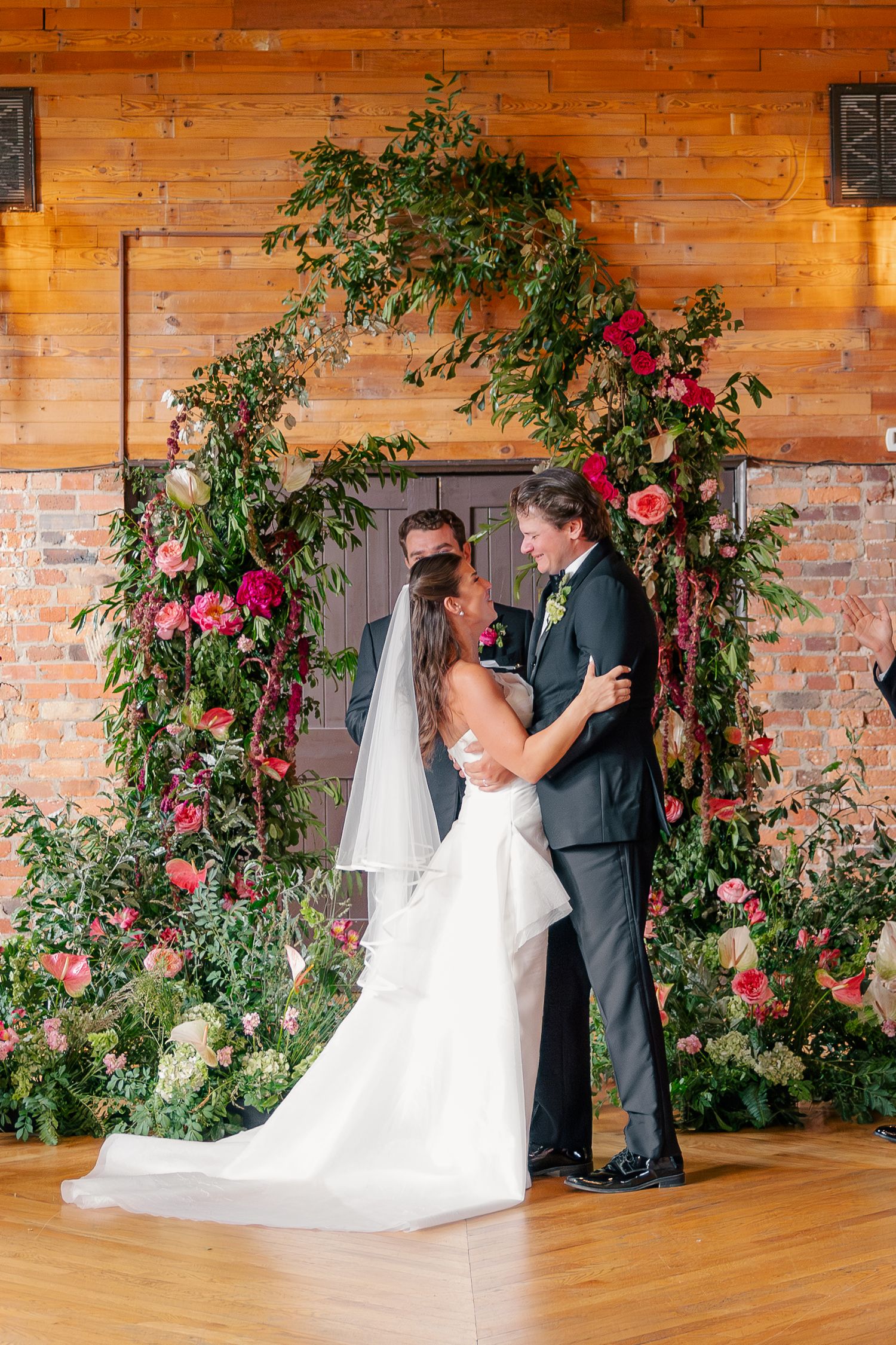 Bride and groom share first kiss under lush floral arch with pink roses and greenery at rustic barn wedding venue.