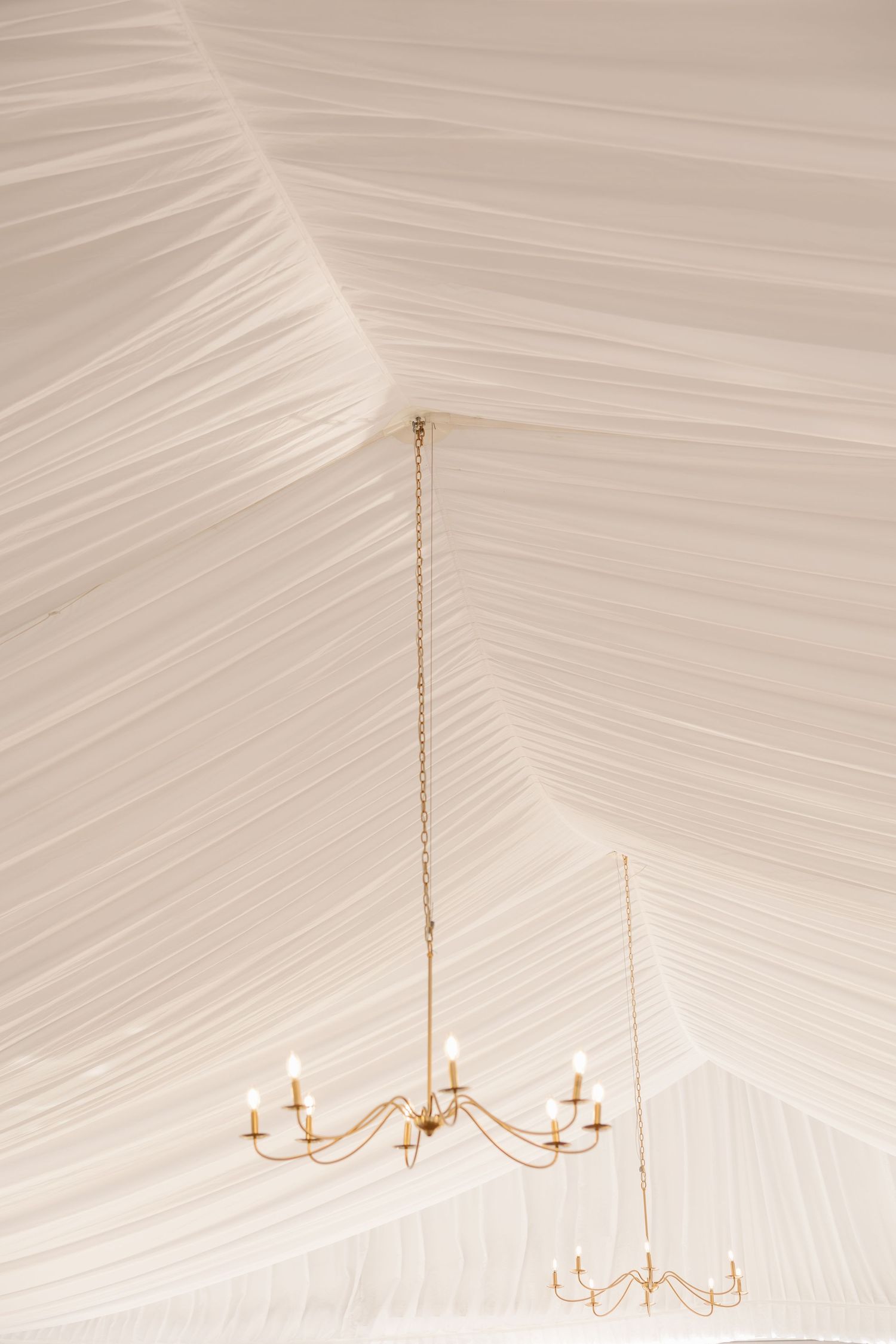 Elegant white draped wedding tent ceiling with gold candelabra chandelier hanging gracefully.