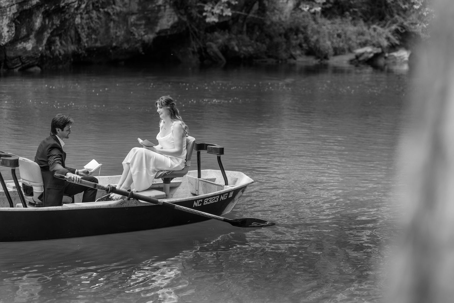 Black and white photo of a couple in a rowboat on a calm river, reading together in elegant wedding attire.