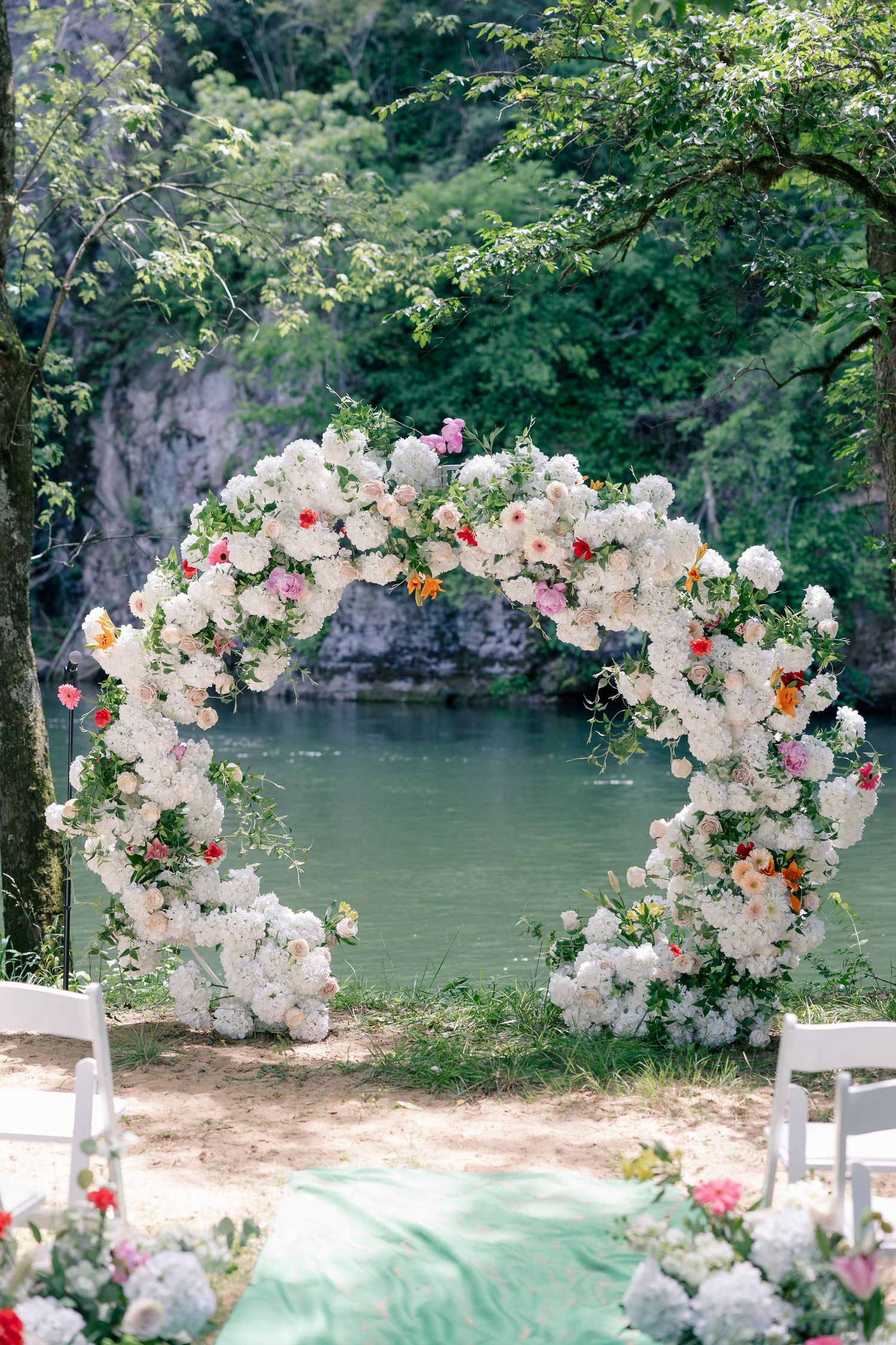 Stunning circular floral arch adorned with white and colorful blooms overlooks a serene turquoise river.