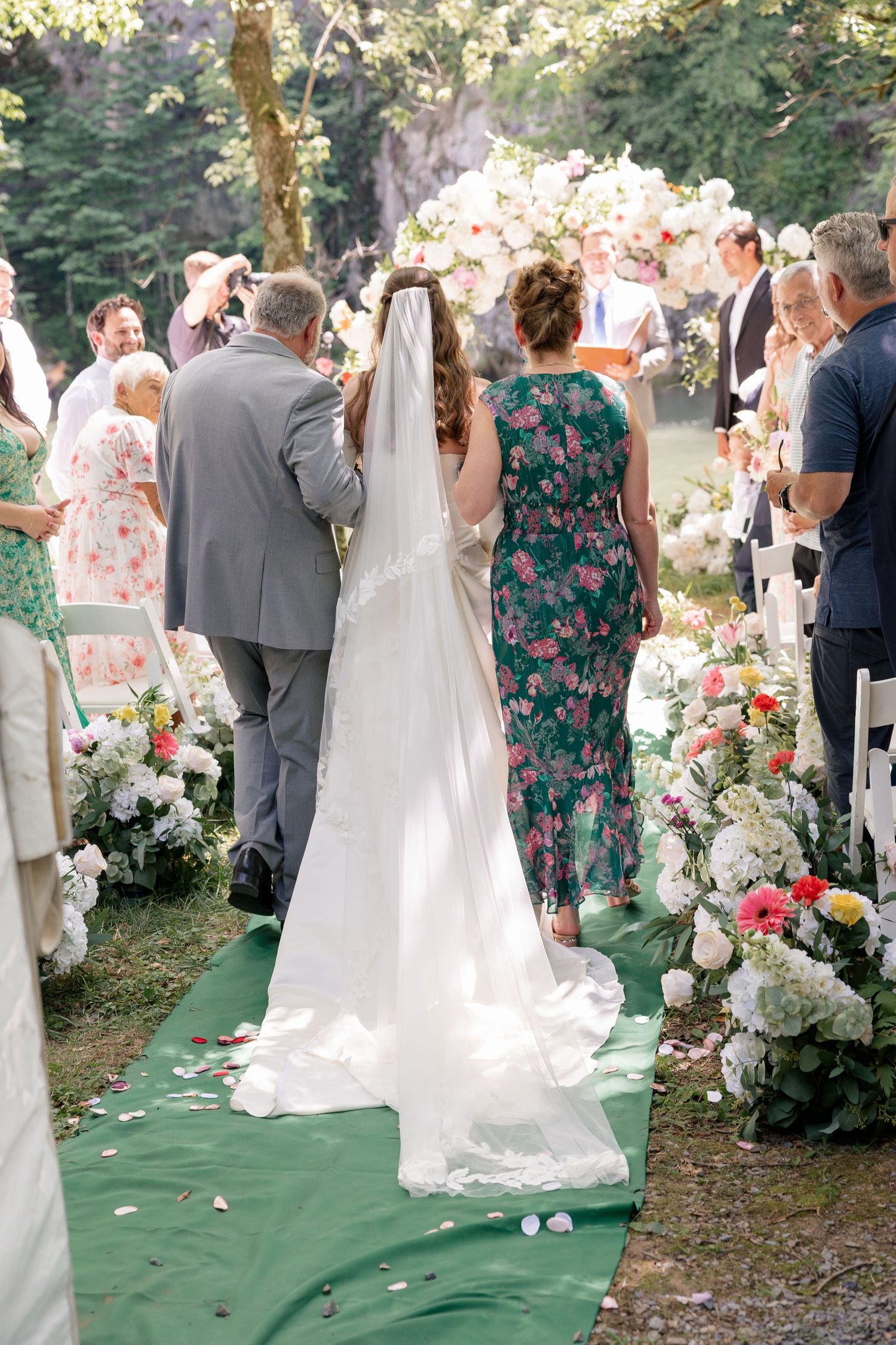 Bride walks down outdoor floral aisle with escort during garden wedding ceremony.