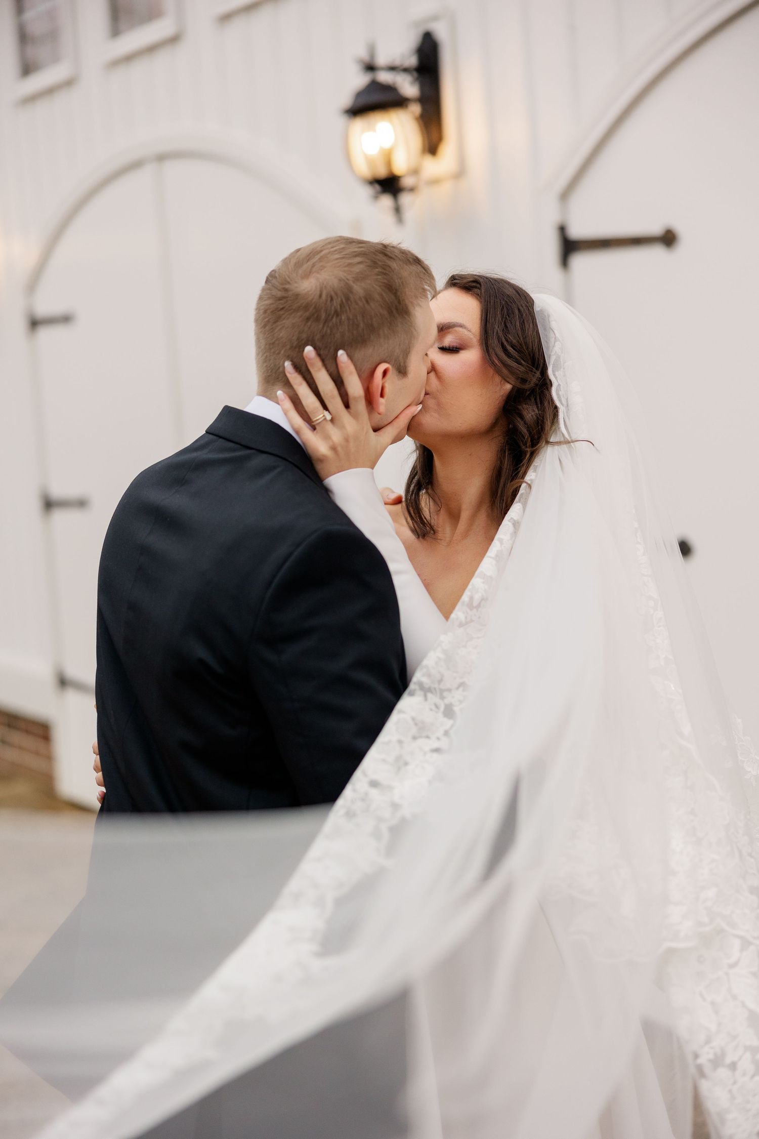 A romantic wedding couple shares a kiss against a white building wall with a glowing lantern in soft lighting.
