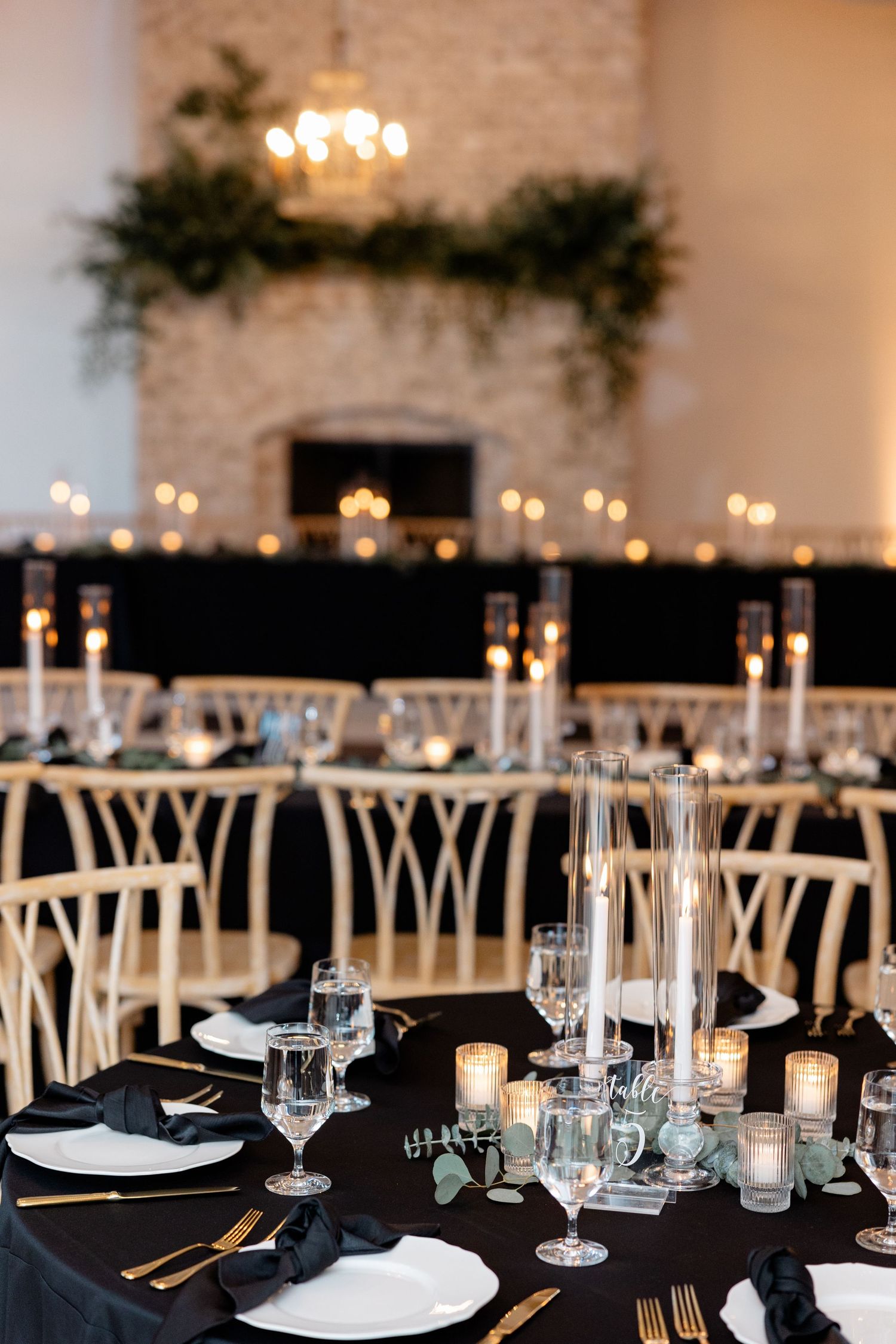 Elegant wedding reception table setup with black linens, candlelight and greenery adorning a white brick fireplace.
