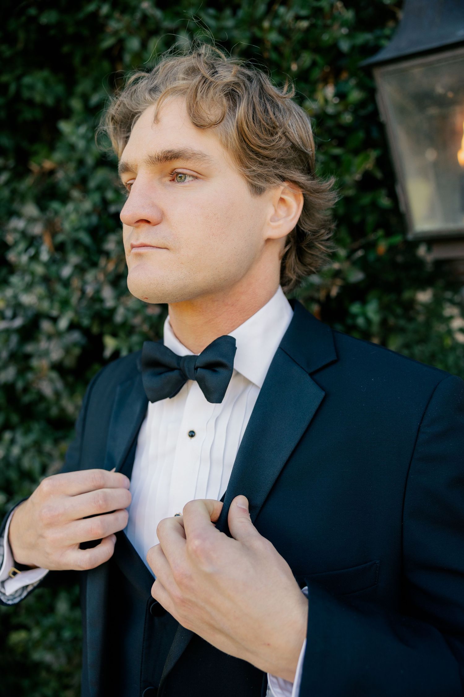 A formal portrait of a person in a black tuxedo and bowtie adjusting their collar against an ivy backdrop.
