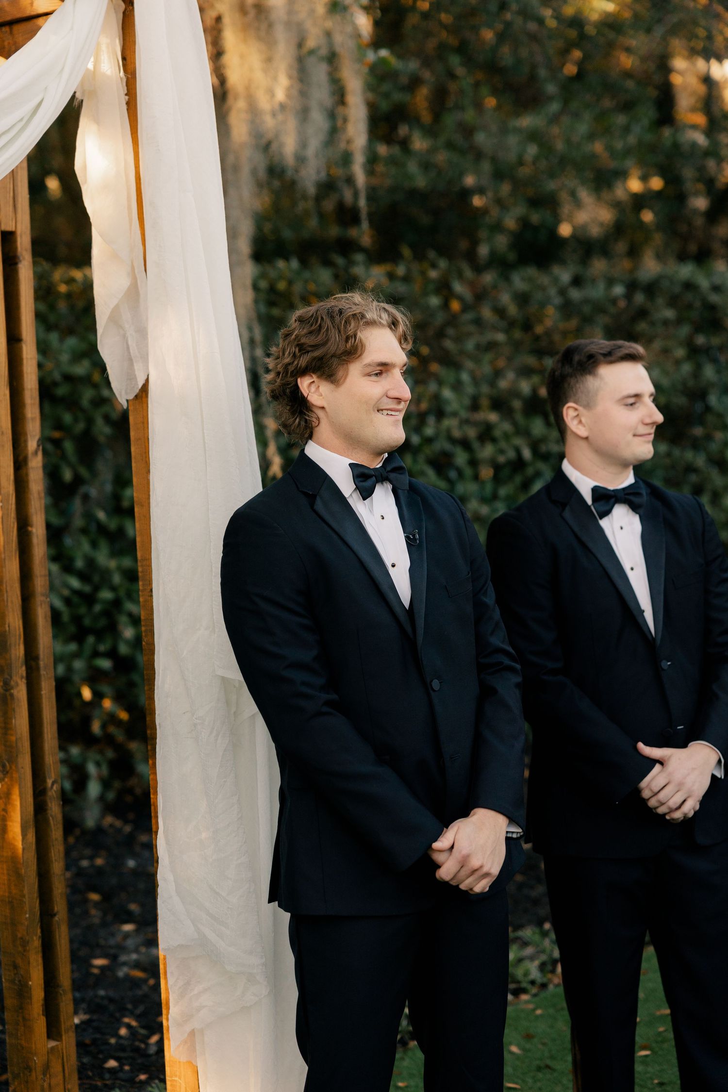 Two groomsmen in classic tuxedos standing at decorated outdoor wedding altar.