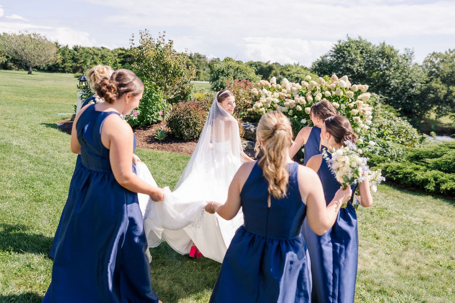 Bridesmaids in navy blue dresses walk with bride in white gown and flowing veil through a garden.