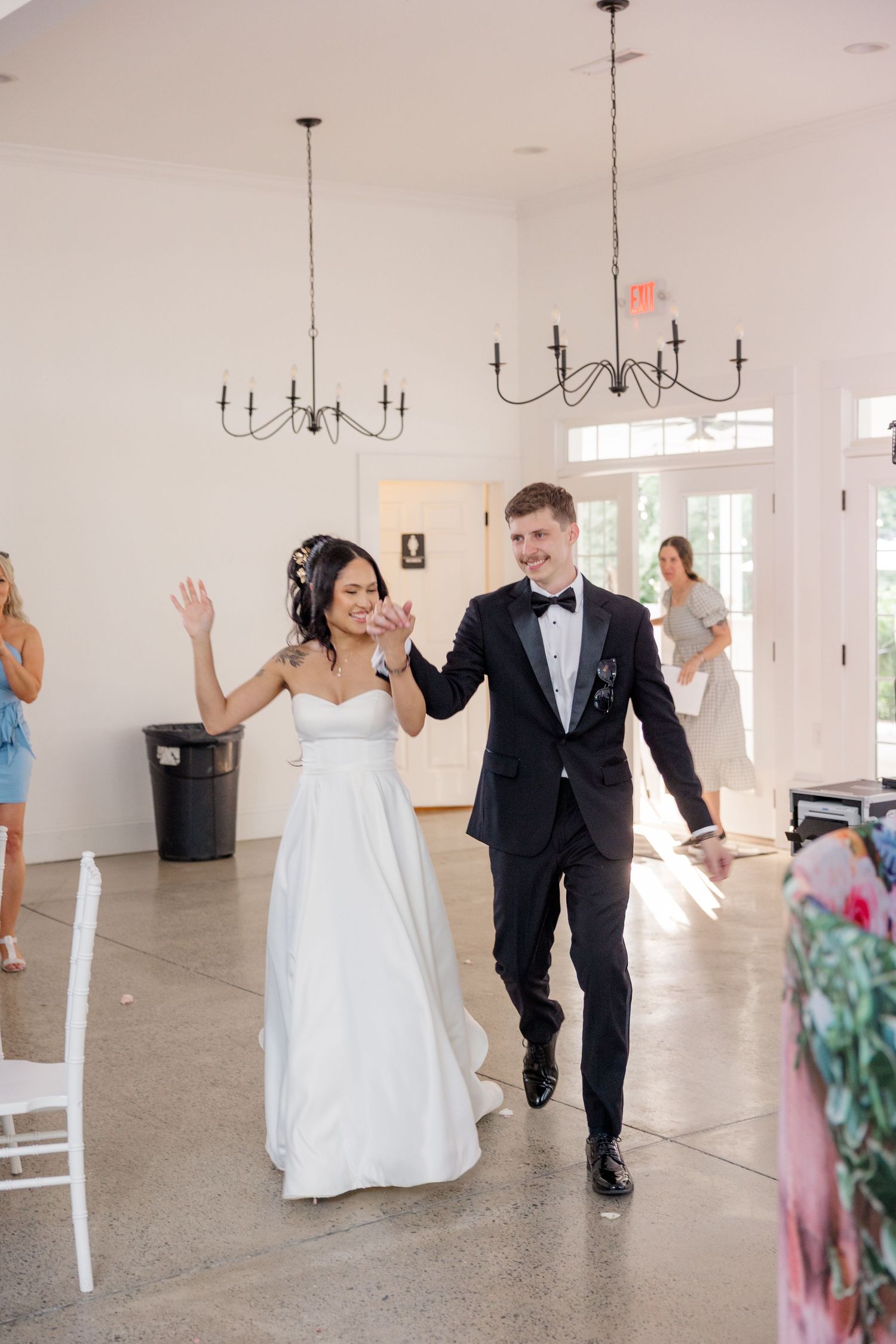Wedding couple makes grand entrance into white reception hall with raised arms in celebration.