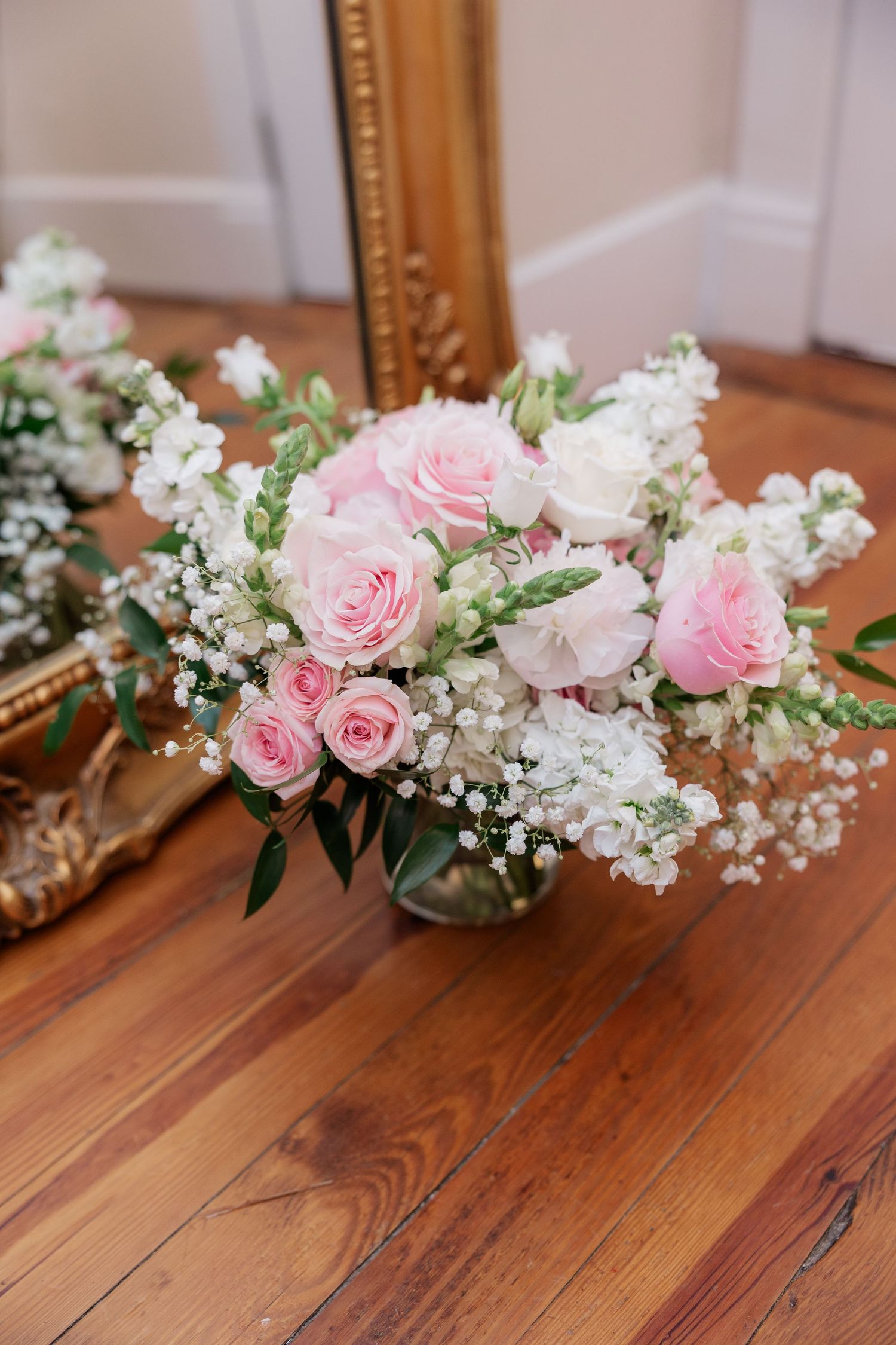 Delicate floral arrangement with pink roses and baby's breath on a wooden surface near a gold-framed mirror.