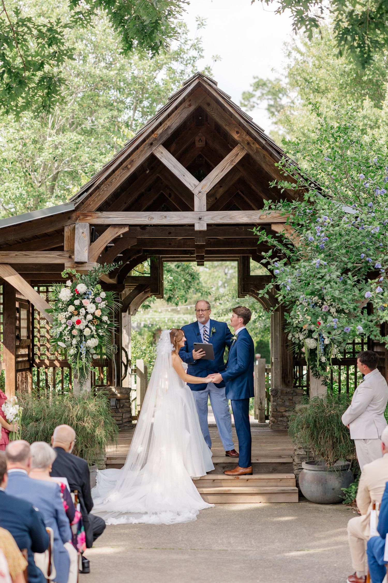 A wedding ceremony takes place under a rustic wooden pavilion surrounded by lush greenery and trees.