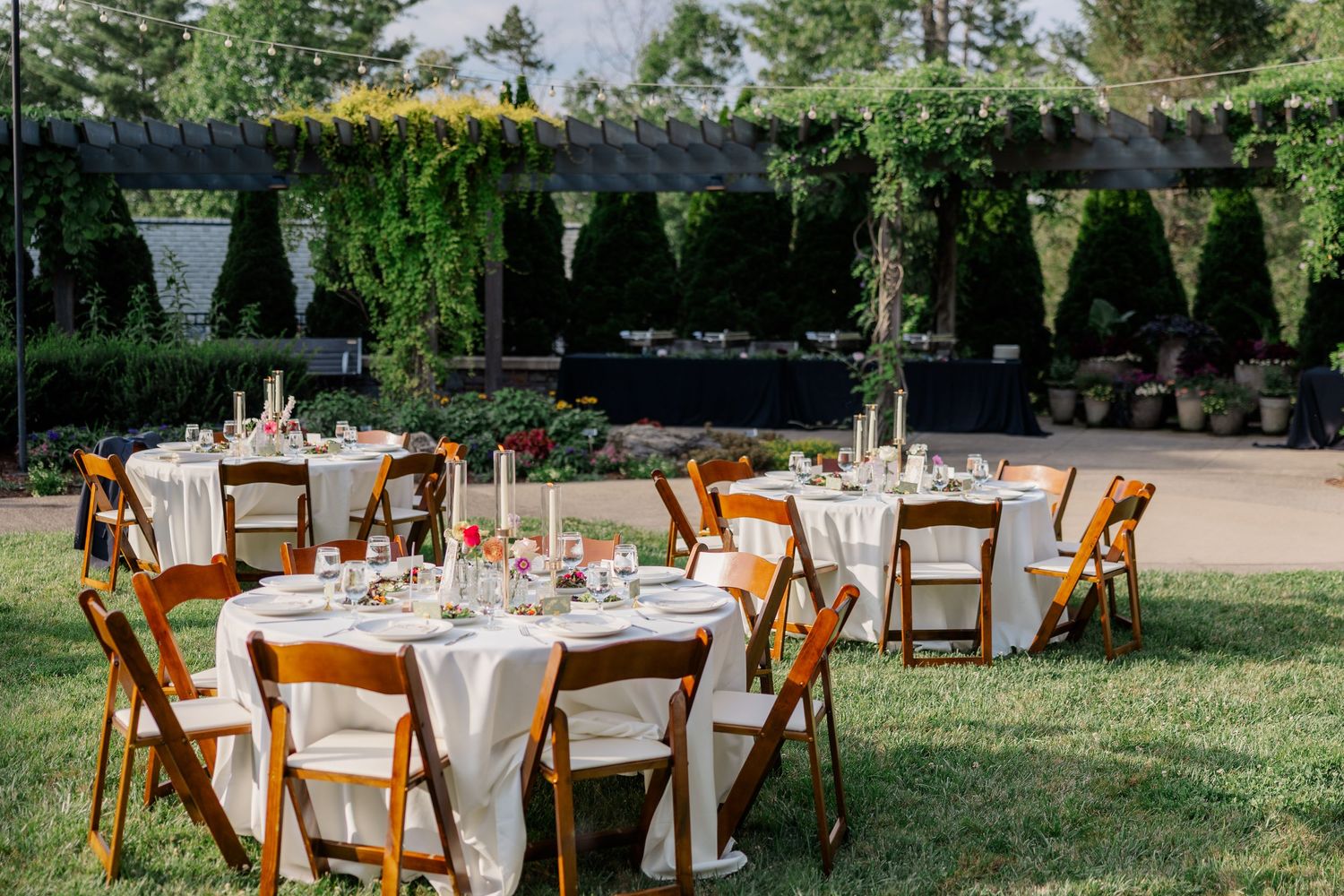 Elegant outdoor wedding reception setup with wooden chairs and round tables on grass.