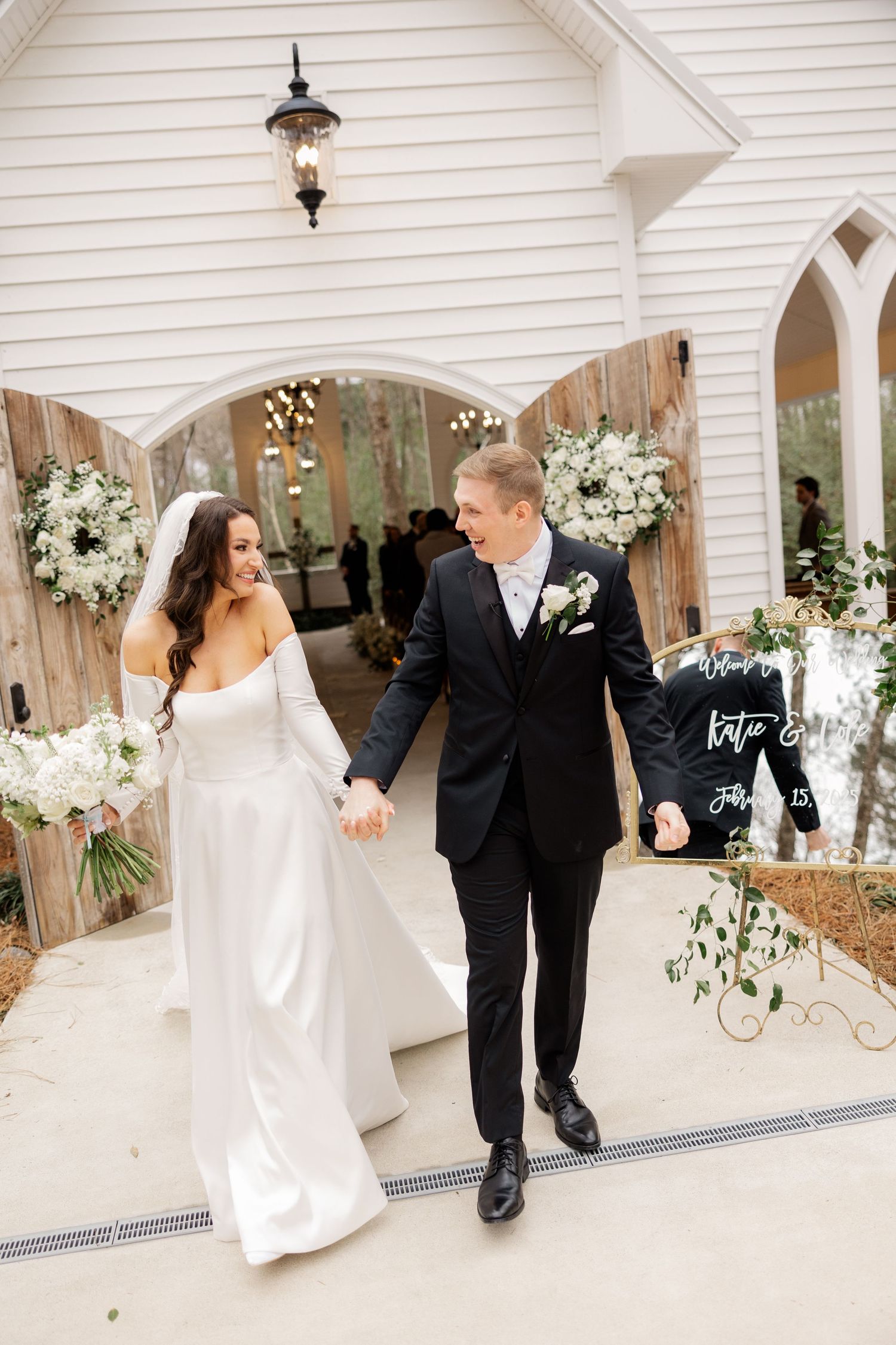 A couple exits a white chapel decorated with floral arrangements and rustic wooden details.