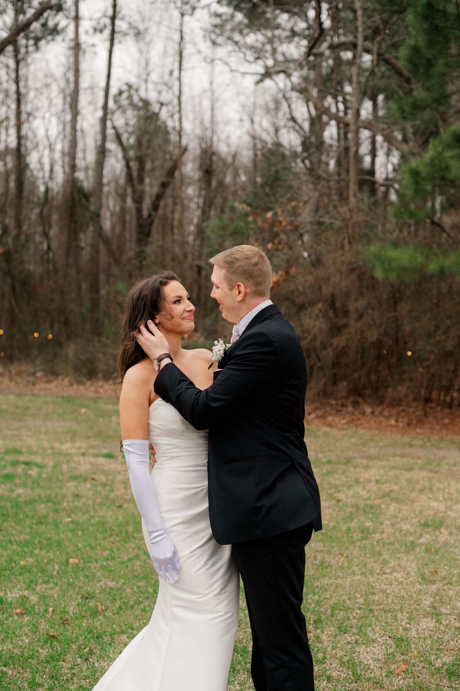 A newlywed couple embraces outdoors in formal attire with trees in the background.