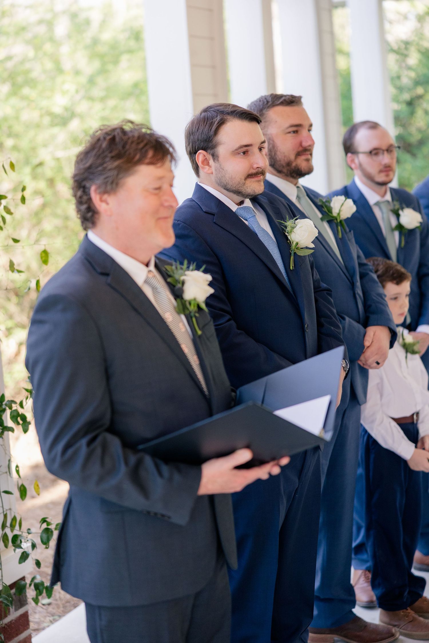 Groomsmen in dark suits stand in line holding papers during a ceremony.