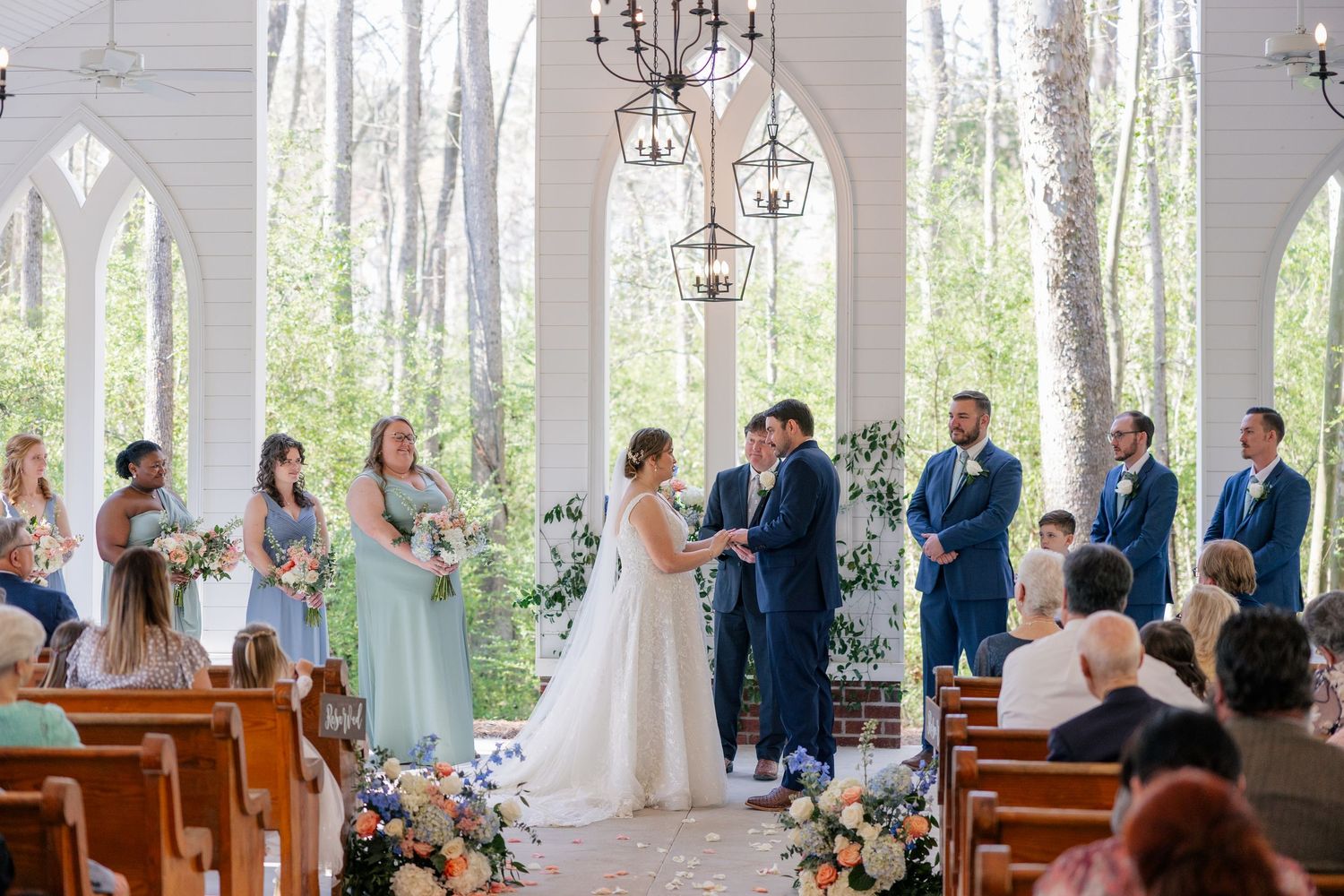 Sun-filled white chapel interior with wooden pews and chandeliers during a wedding ceremony.