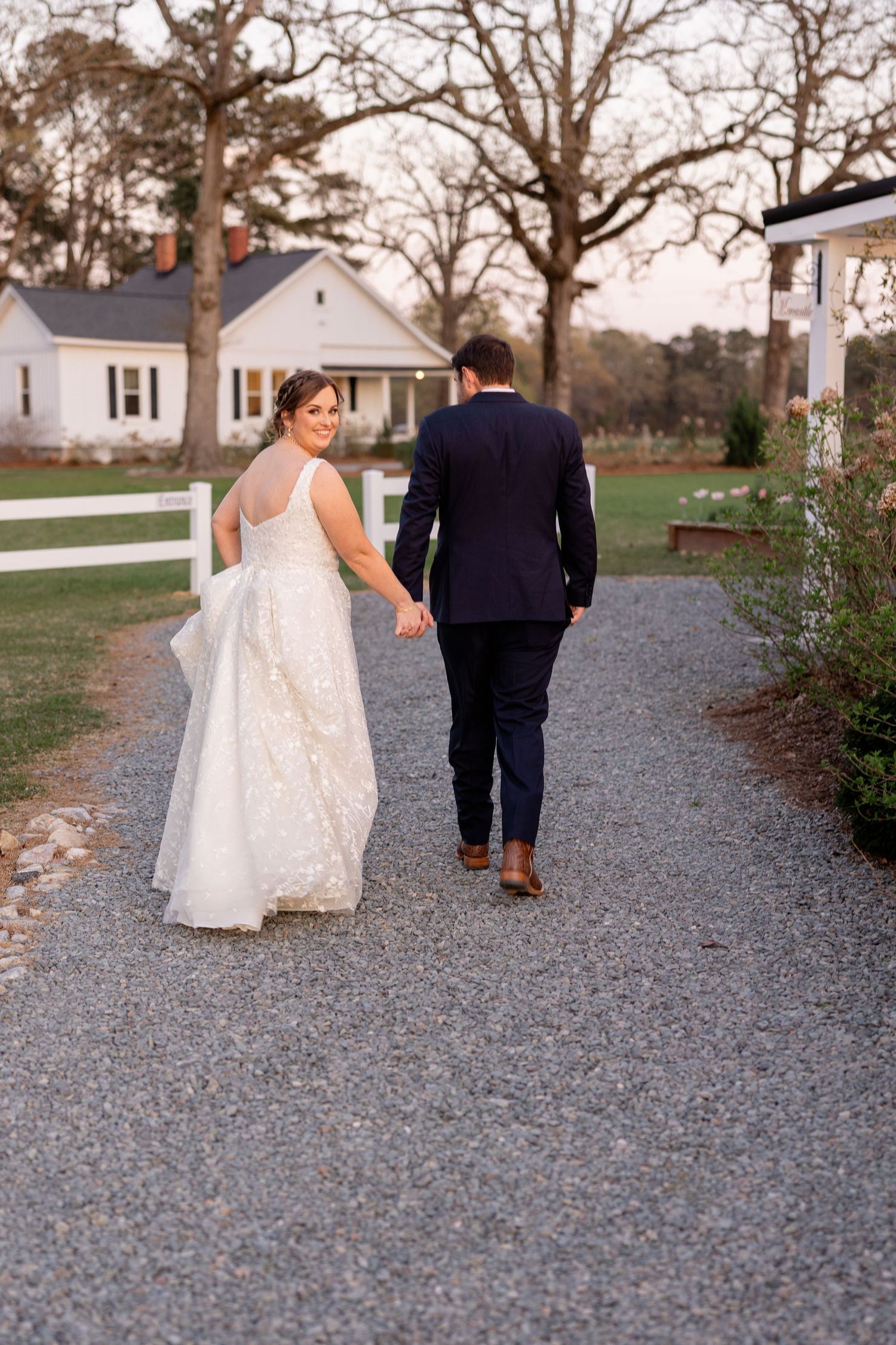 Bride and groom walking down a gravel path with white house in background.